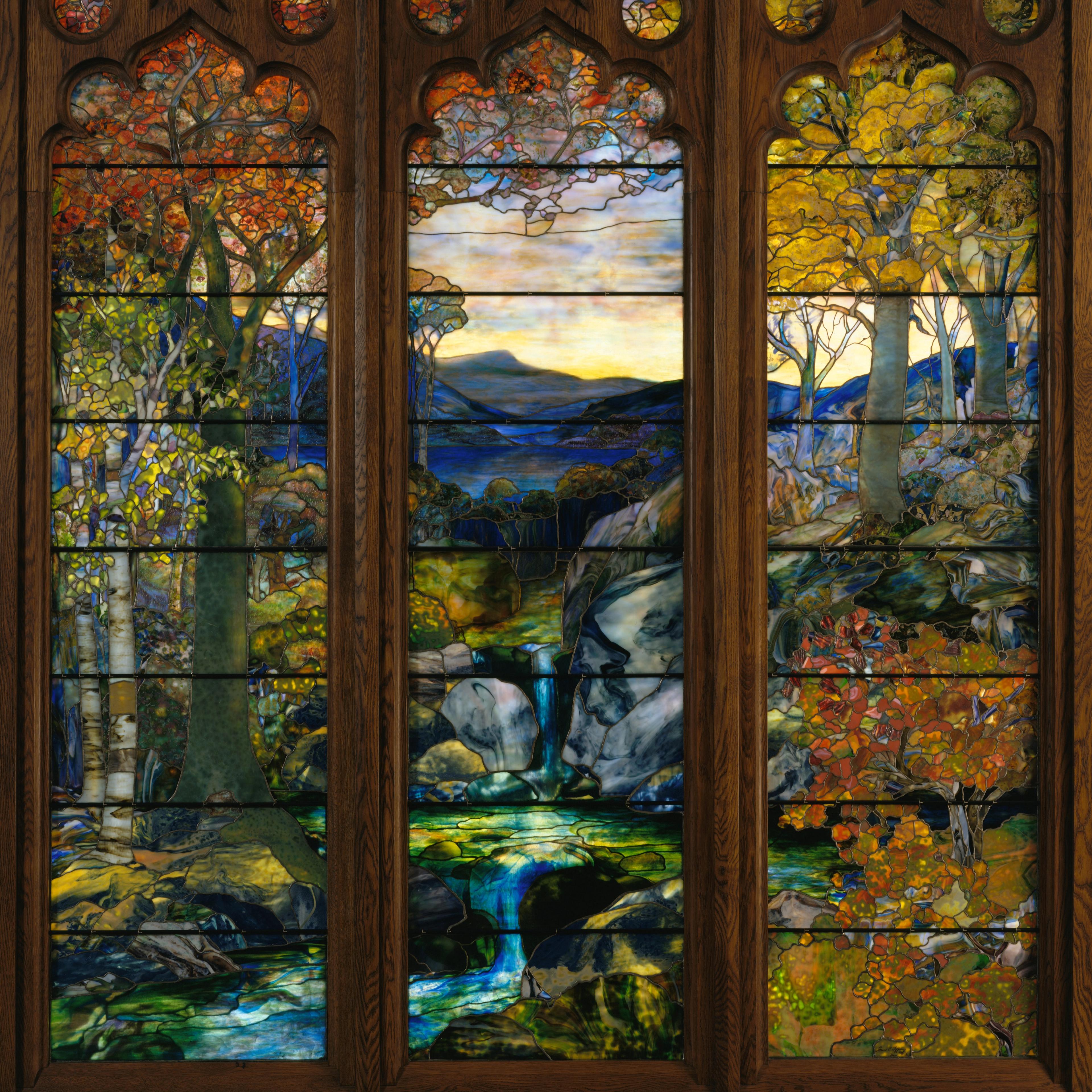 A tall, colorful stained-glass window depicting a forested landscape with flowing water, autumnal trees, and distant mountains.