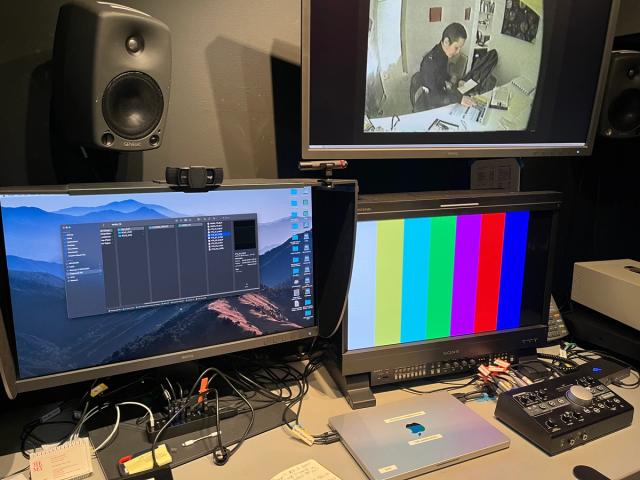 A workspace with three monitors: one showing a file directory, one displaying color bars, and one playing a video of a person sitting at a desk, surrounded by audio and video equipment.