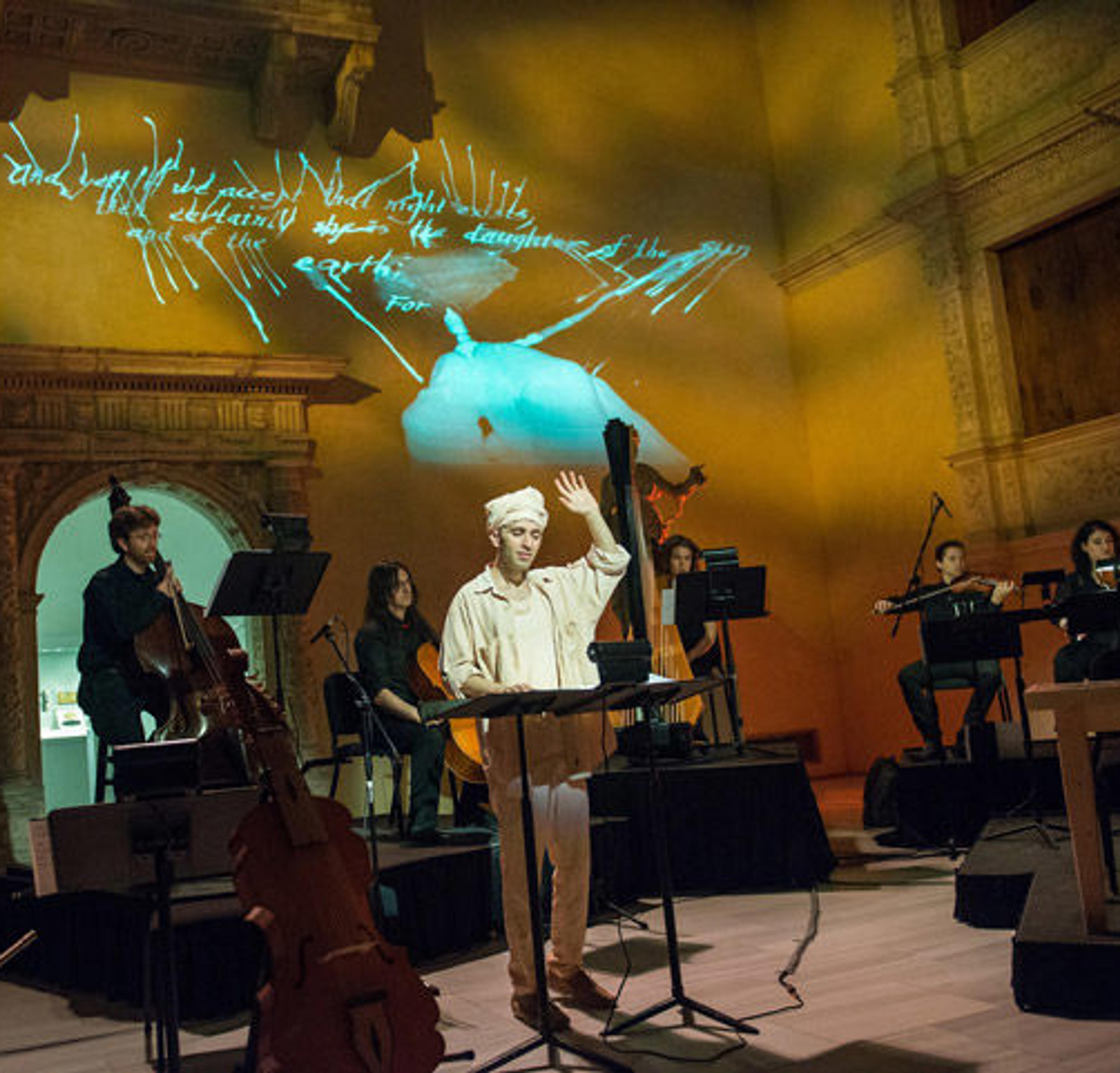 A singer wearing white clothing and a turban performs in front of a chamber orchestra while surrounded by European sculpture