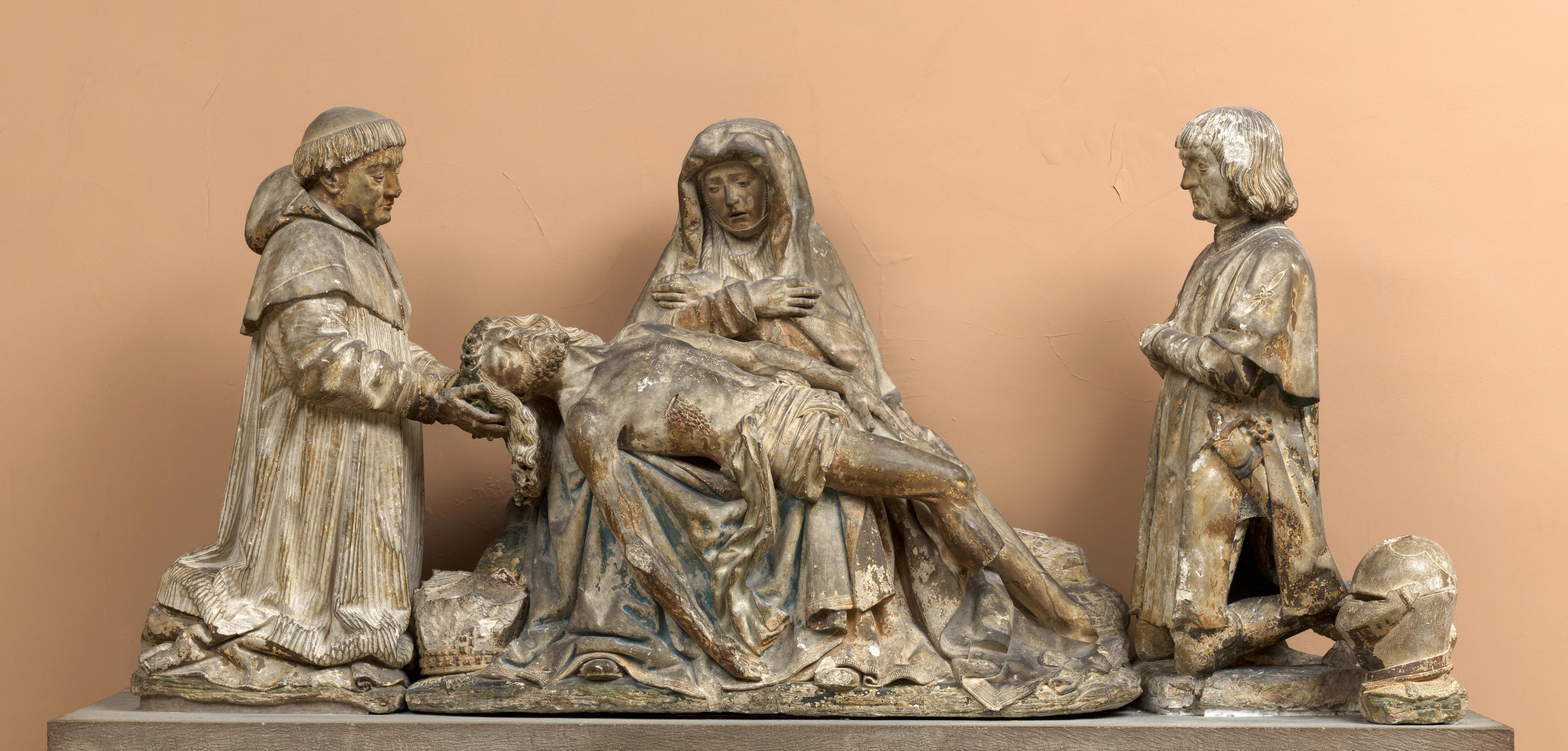 Sculpture showing two men kneeling on either side of a cloaked Virgin Mary with Christ stretched across her lap. The sculpture appears heavily stained.