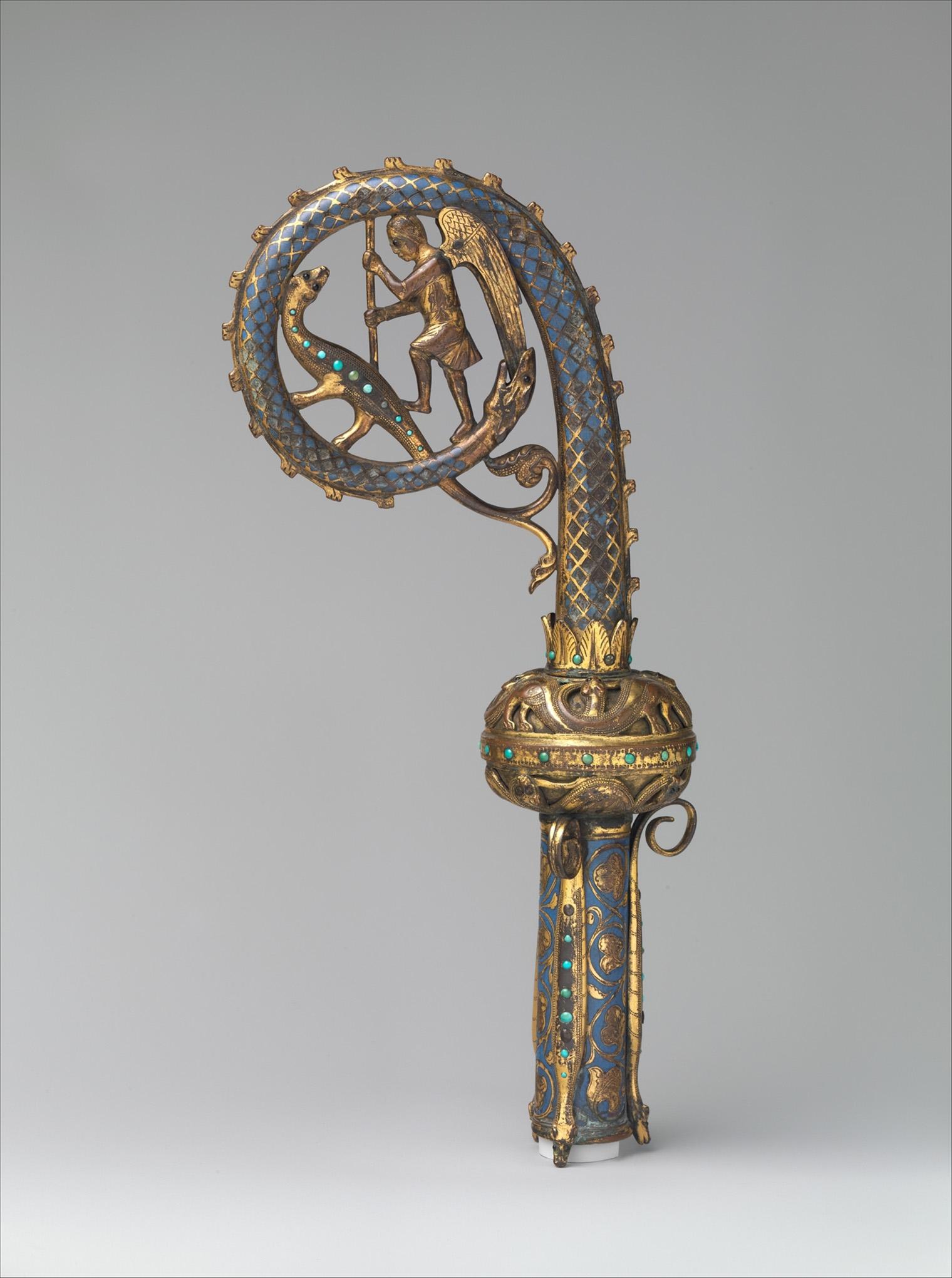 Ornate gold crozier with an intricate spiral top, featuring an angel and a deer, adorned with blue enamel and green gemstones.