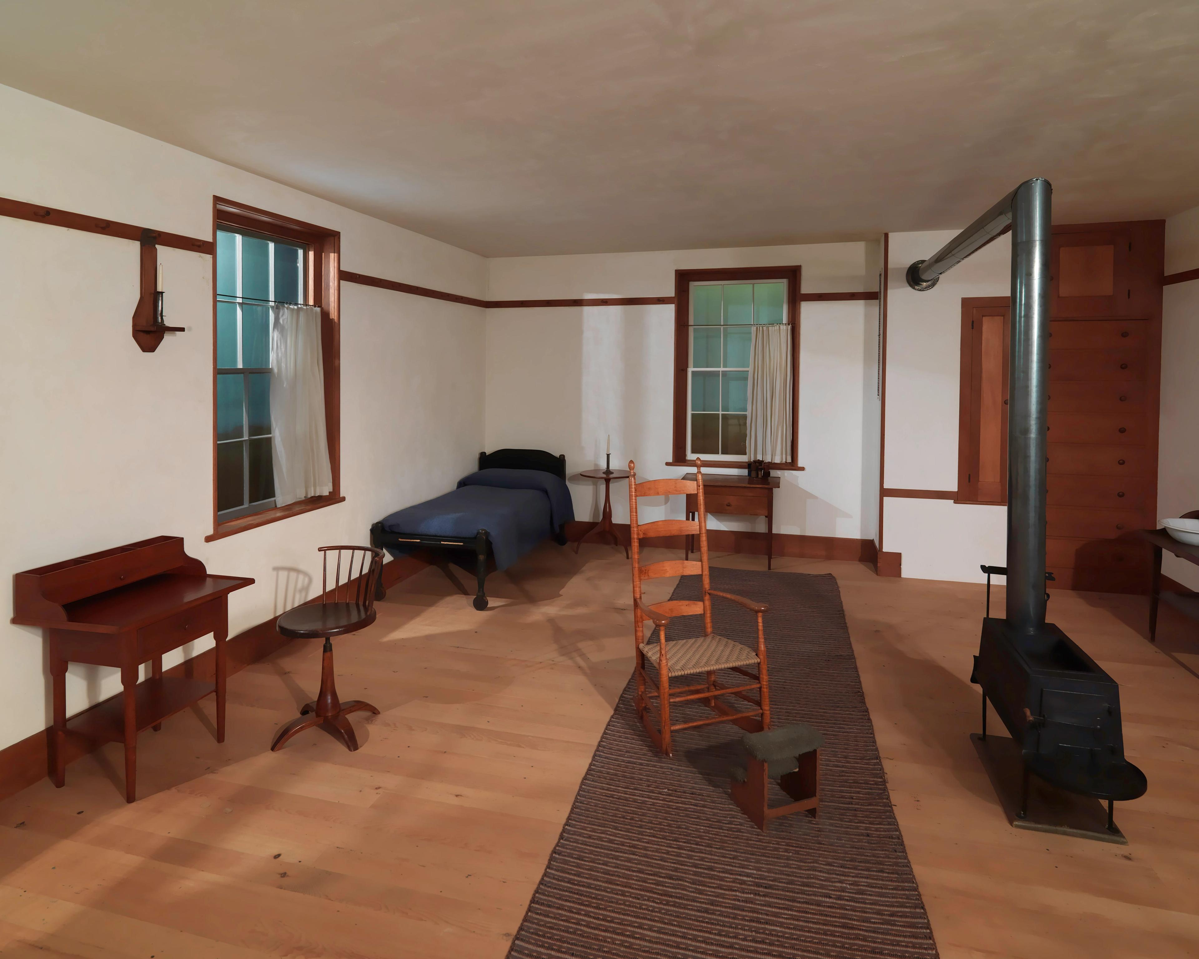 A simple, sparsely-furnished room with wooden floors, a single bed, wooden chairs and tables, and a tall black stove.