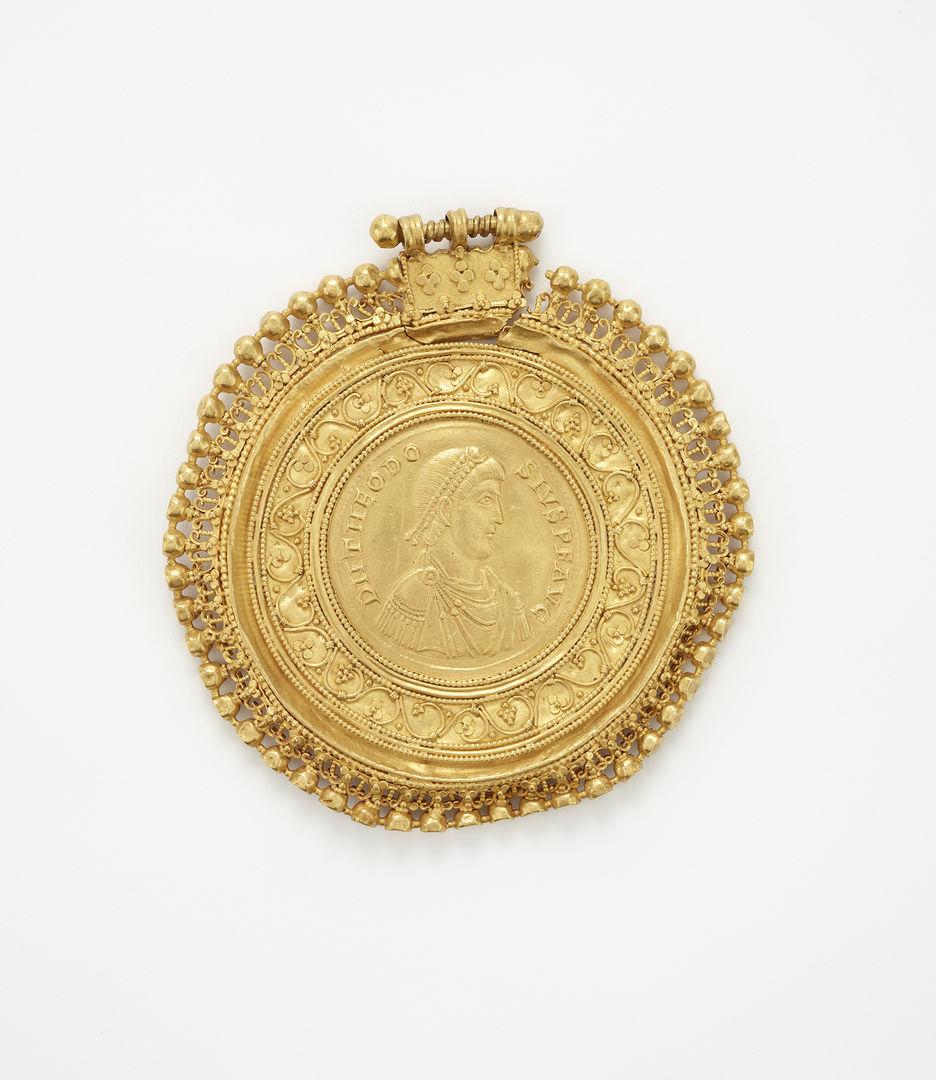 A gold coin of Theodosius I at the center looking out to the right. He is bordered by elaborate decorative elements around the coin.