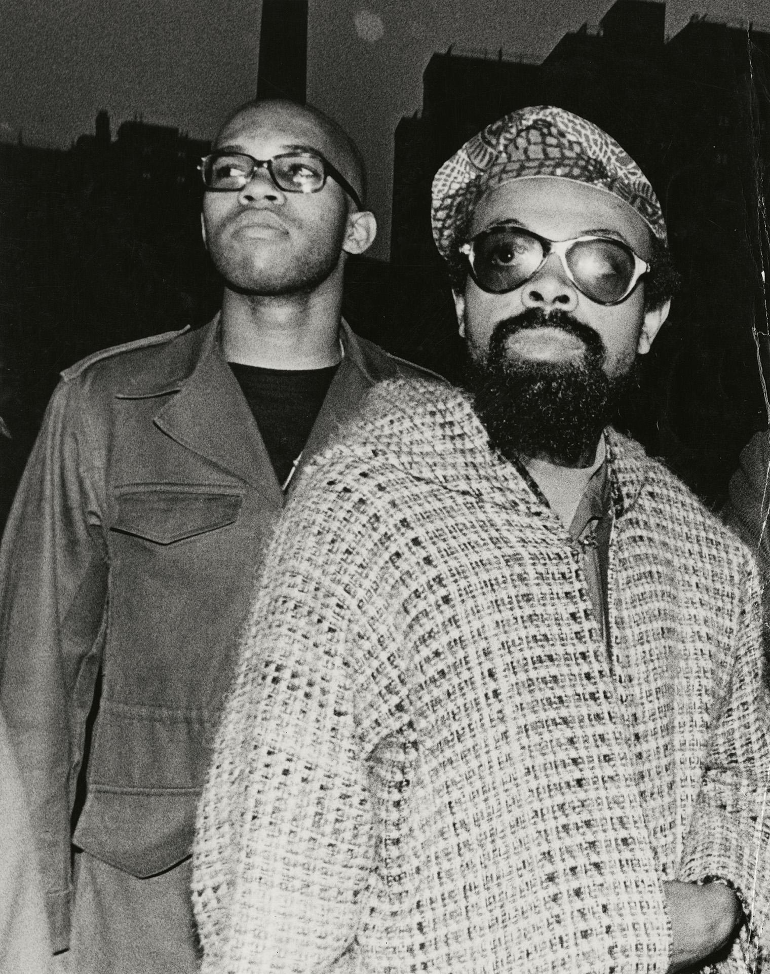 Two men stand together in a black-and-white photo. The man in front wears sunglasses, a patterned coat, and a beret. The man behind wears glasses and a jacket, exuding a serious, contemplative mood. Dark buildings silhouette the background.