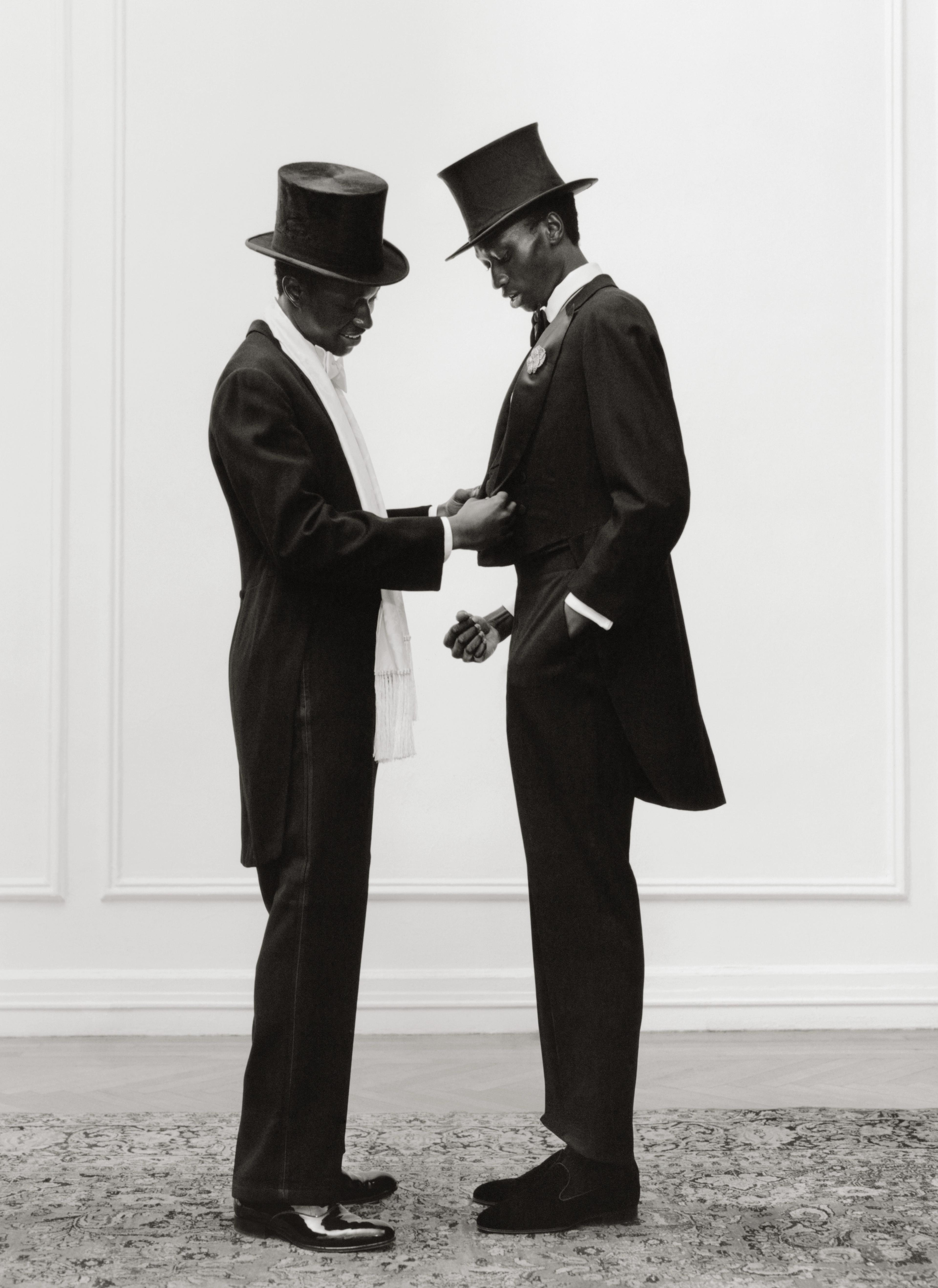 Black and white photograph of two men in suits and top hats. The man on the left adjusts the suit of the other.