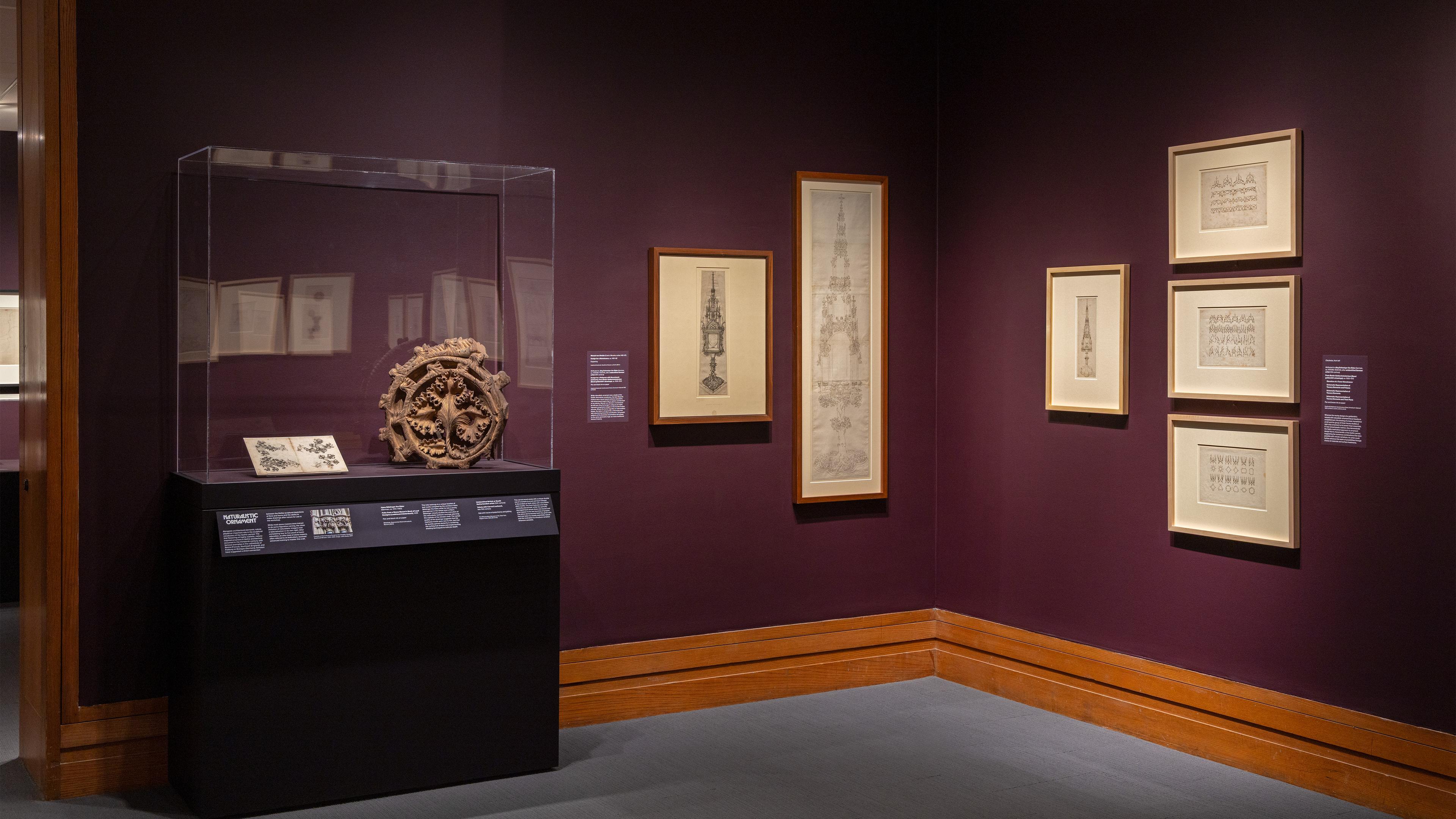 A dimly lit museum exhibit with framed sketches on deep purple walls. A glass case displays an intricate circular sculpture.