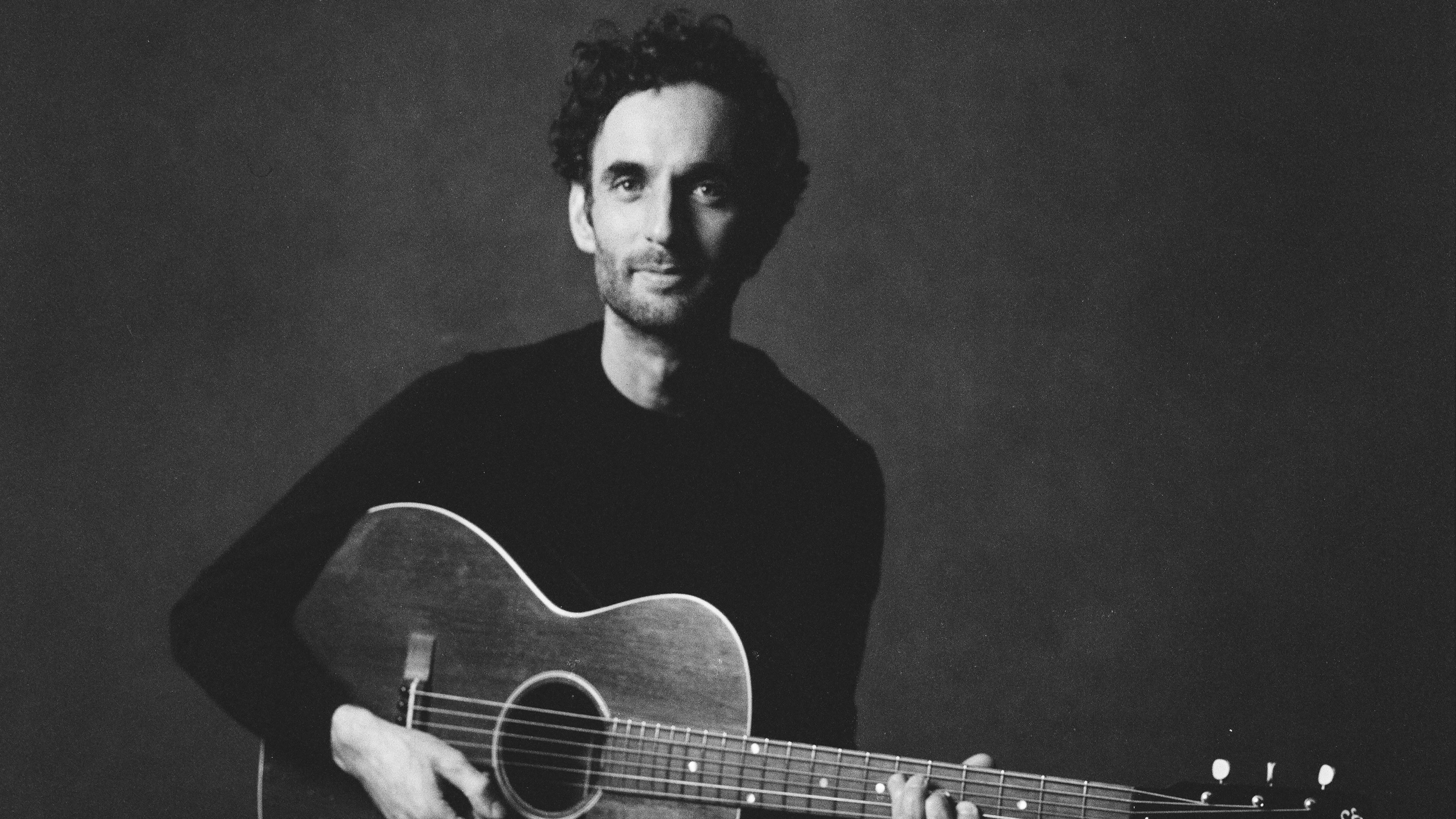 A man in a black sweater holds an acoustic guitar.