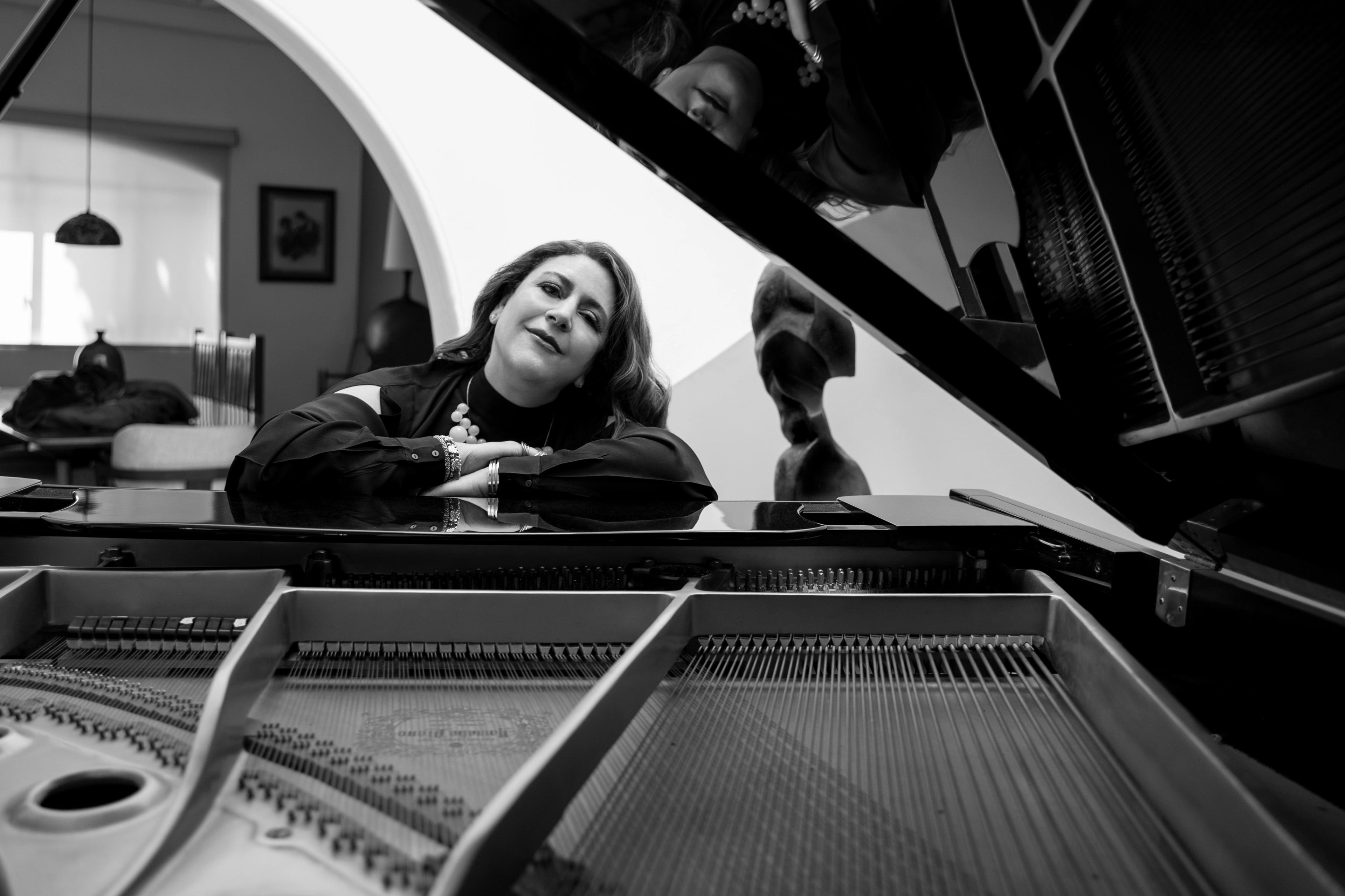 Gabriela Ortiz, softly smiling, leans with her arms crossed over the top of a piano.