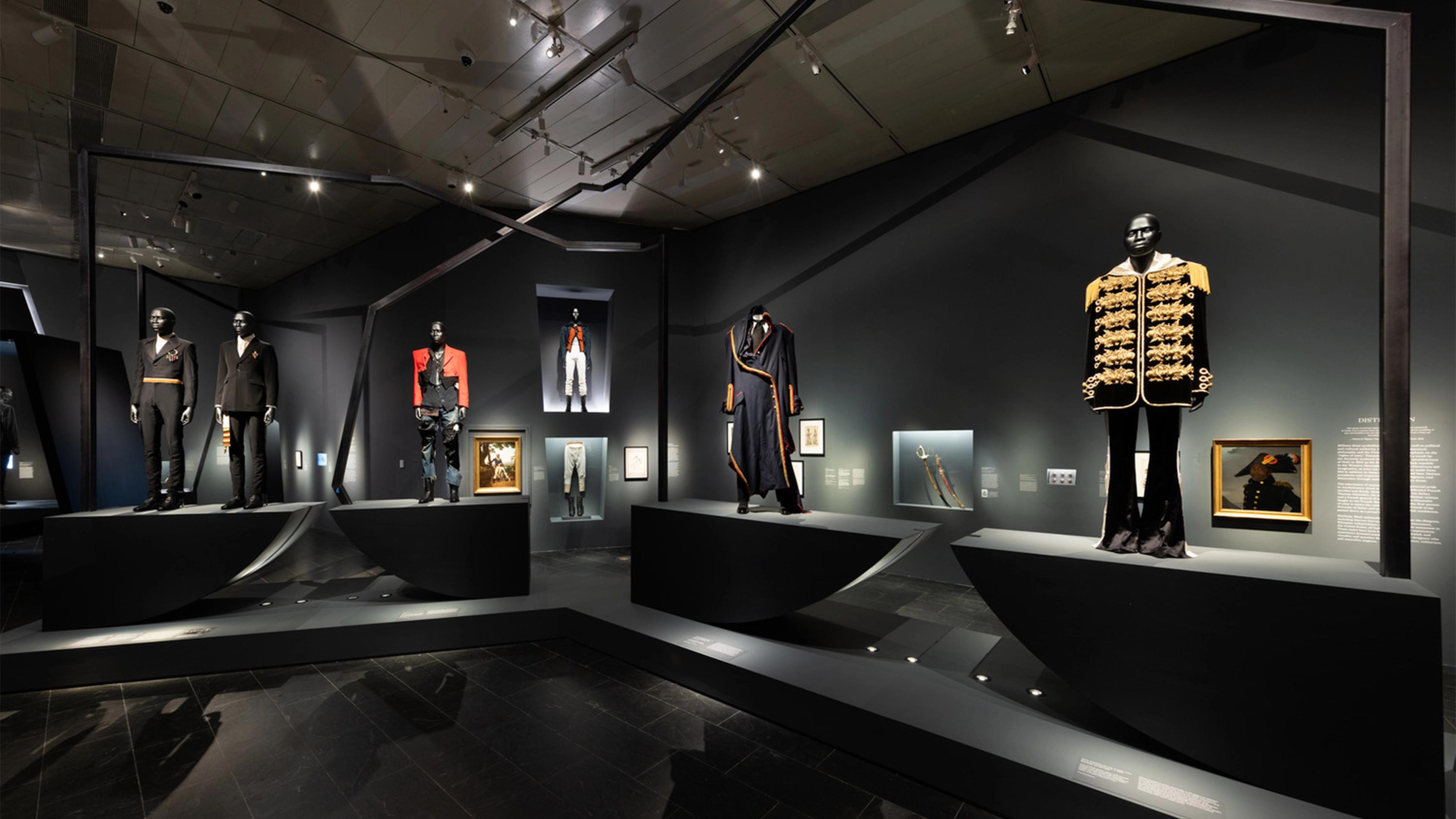 Museum exhibit showcasing historical and military-inspired fashion. Mannequins display ornate, dark uniforms with bright accents, set against a dimly lit backdrop.
