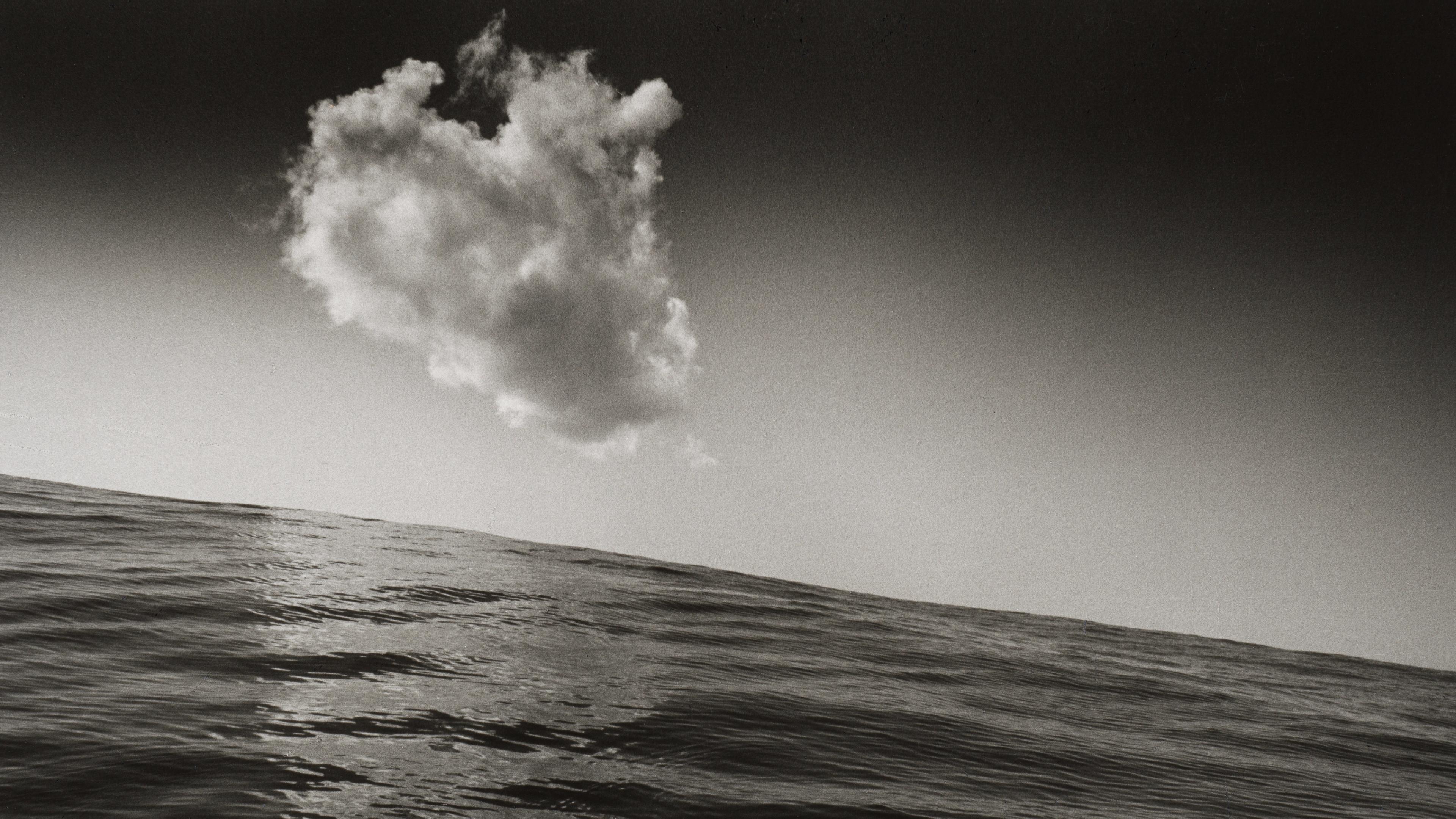 Black-and-white seascape with a large, billowing cloud above a slanted ocean horizon; textured waves and dramatic contrast .