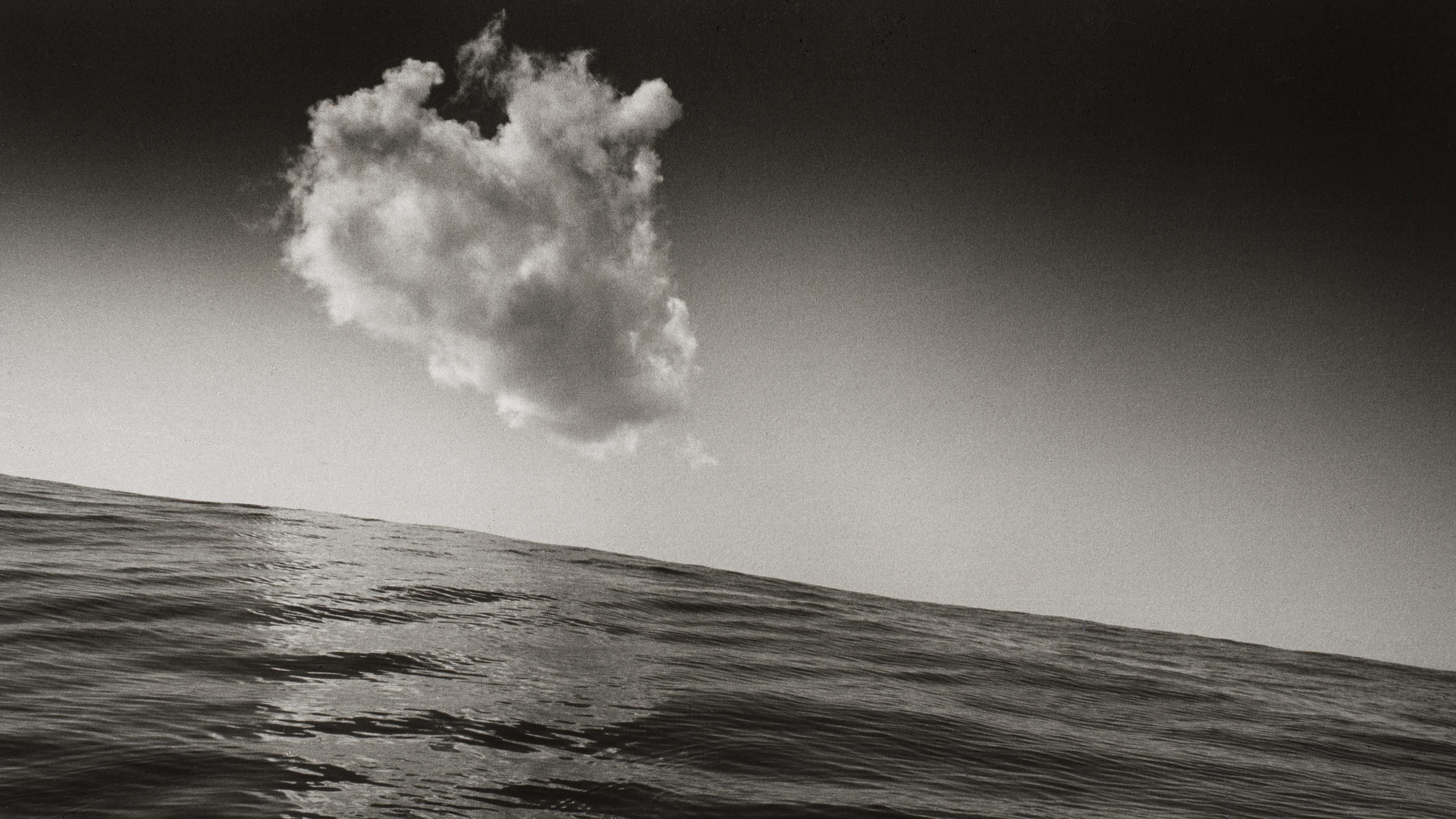 Black-and-white seascape with a large, billowing cloud above a slanted ocean horizon; textured waves and dramatic contrast .