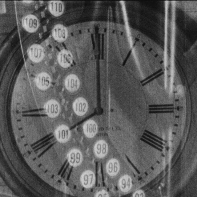 A black-and-white still from the 1928 film Behind the Scenes: The Working Side of the Museum, which depicts the superimposed image of an old-fashioned time-card machine over the face of a wall clock