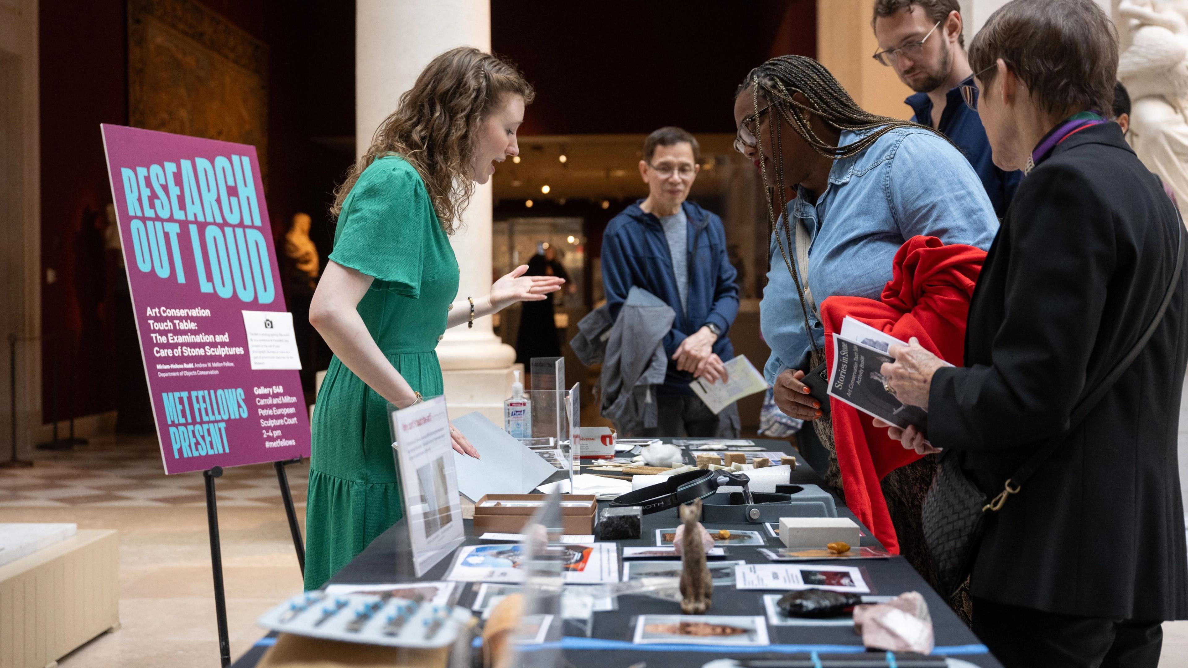 A wide shot in a brightly lit art museum shows a woman in a light green dress leading a presentation at a table. A bright purple and blue poster that reads 'Research Out Loud Met Fellows Present' stands on a black easel behind her. She gestures while speaking to four visitors. The table is covered with pamphlets and objects related to art conservation.