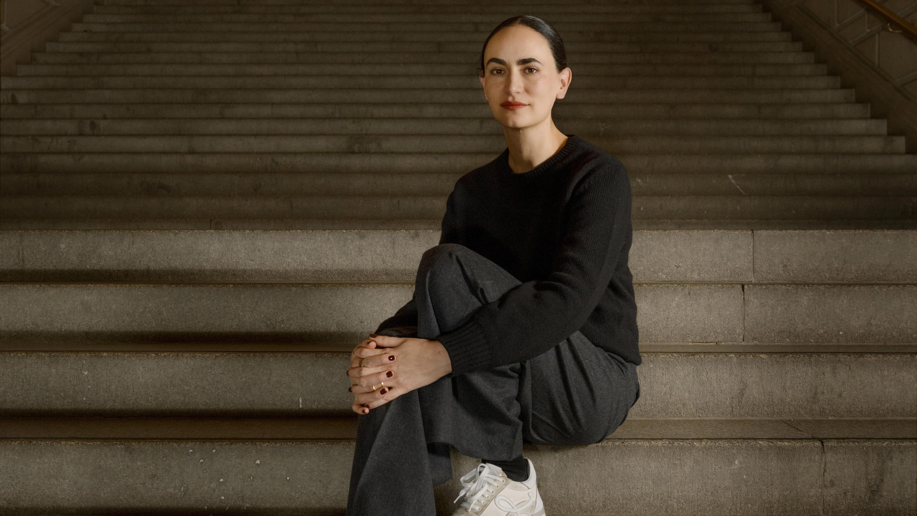 A person with dark hair in a black sweater and gray pants sits on stone steps