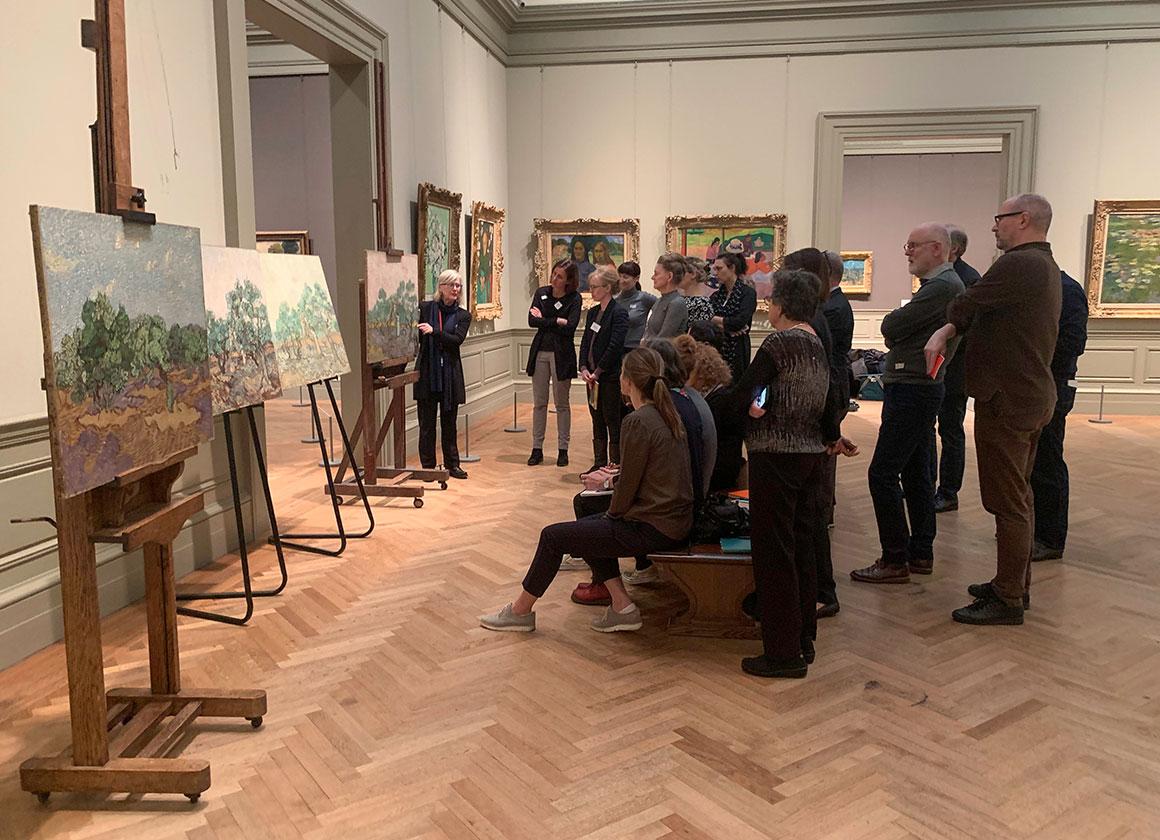 A large group of men and women looking at four paintings on easels stationed in a Museum gallery. A conservator stands at the left of one of the paintings making a presentation to the crowd.