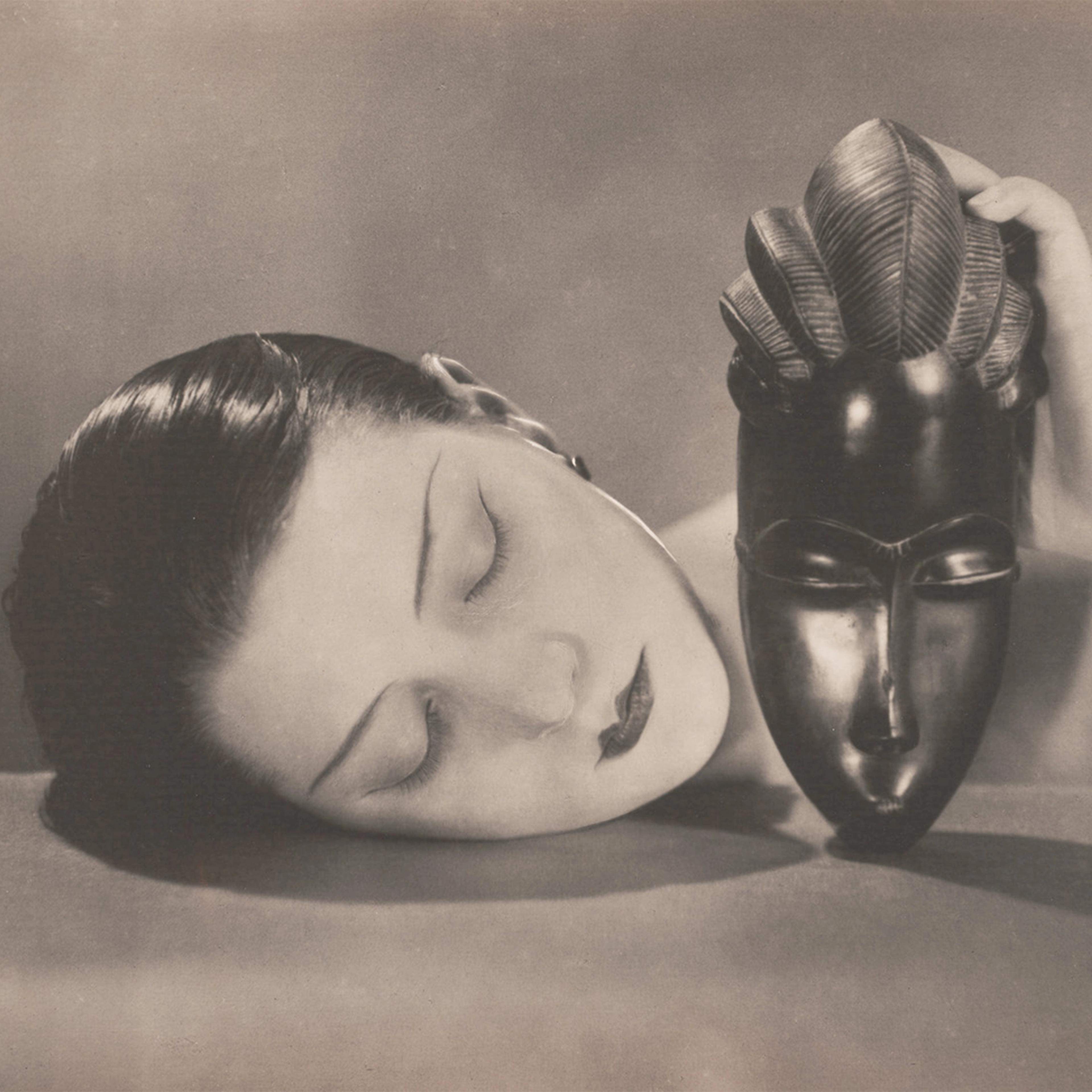A woman with closed eyes rests her head on a surface, holding an ornate African mask. The sepia-toned image conveys a serene and introspective mood.
