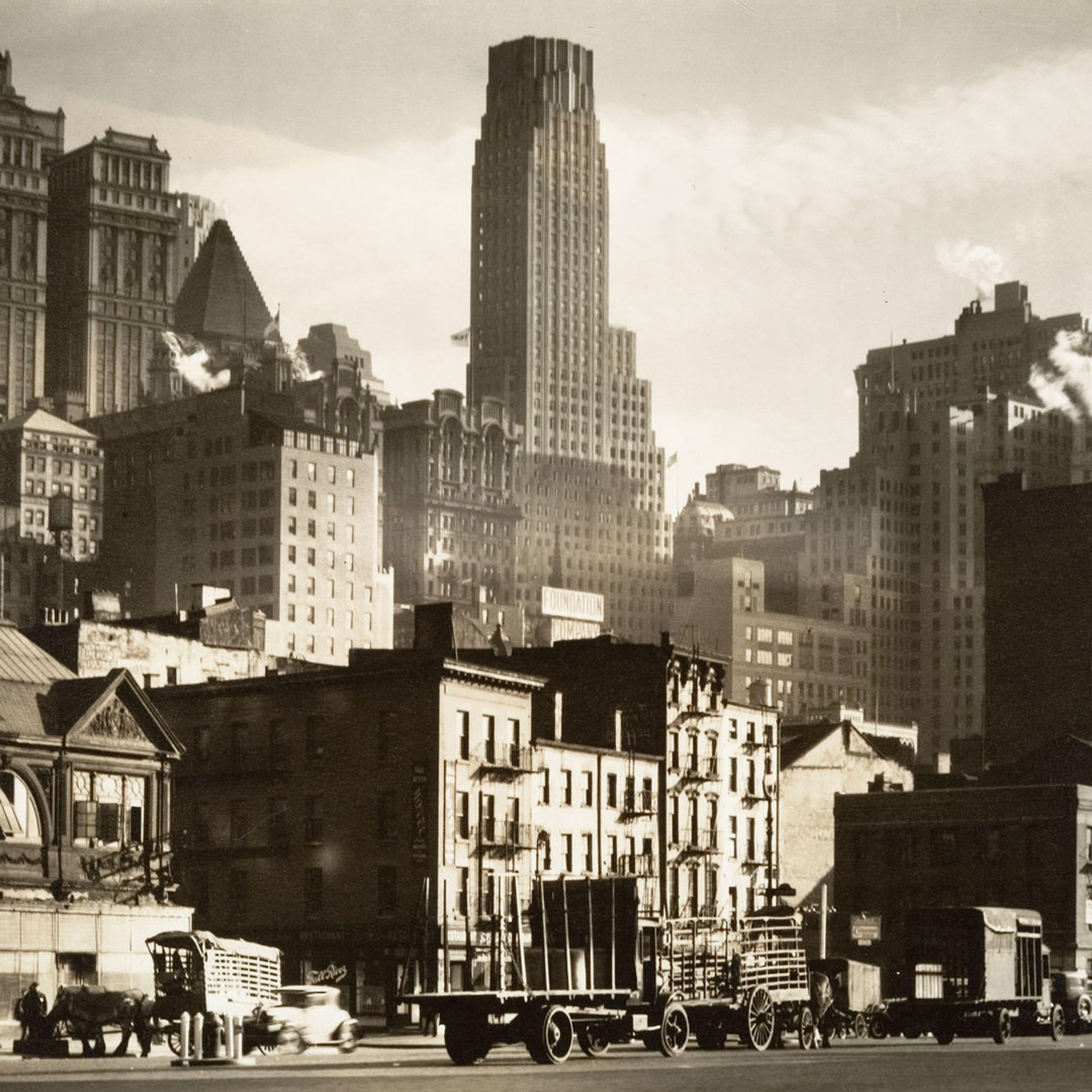 Sepia-tone photograph of old-timey wagon trucks and cars traveling up and down West Street in New York City set against a backdrop of multi-story apartments and midsize skyscrapers.