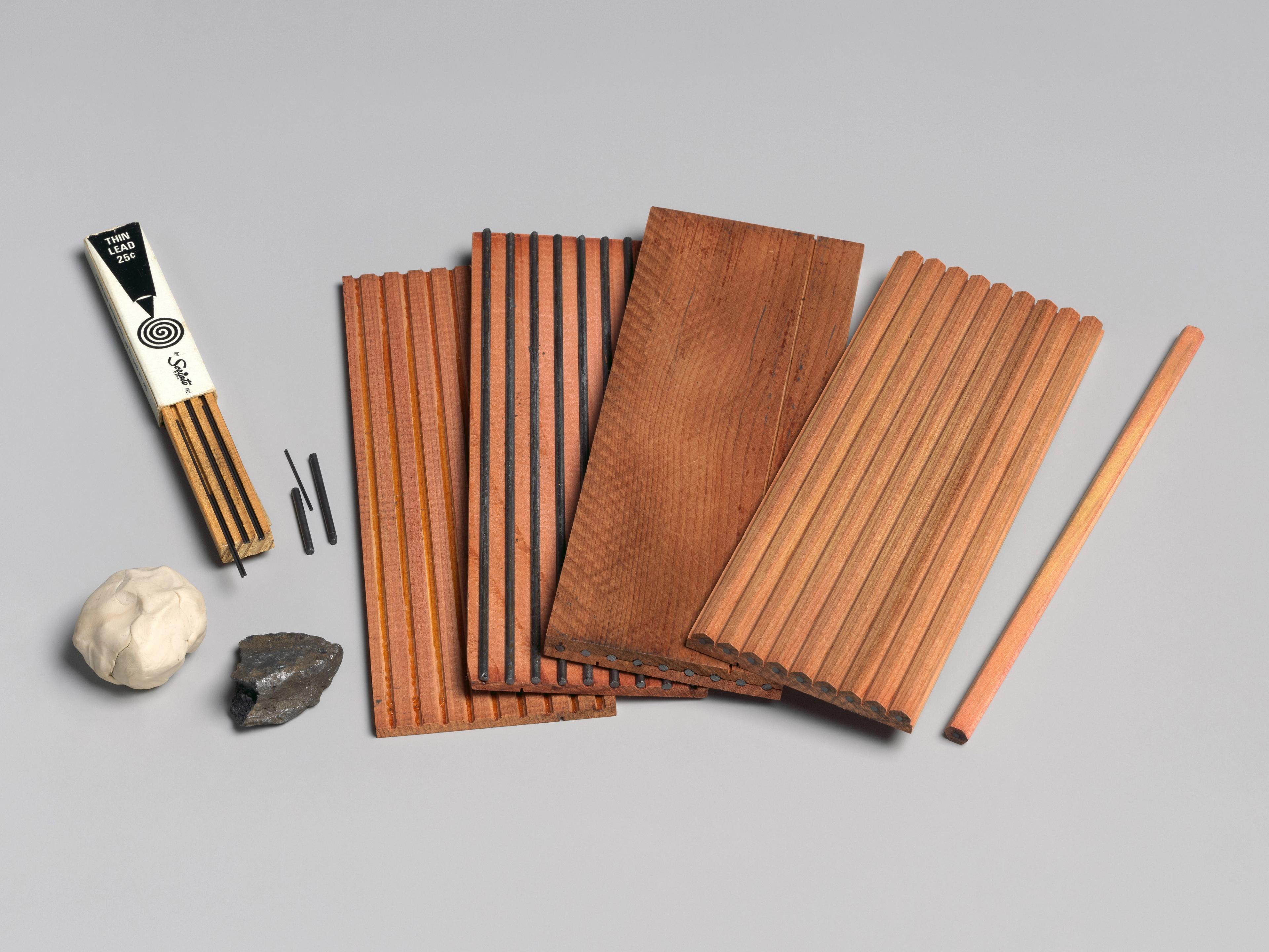 An arrangement of materials used to make pencils, showcased in multiple stages of the process. To the left, a kneading eraser, raw graphite, and a small container of thin lead.