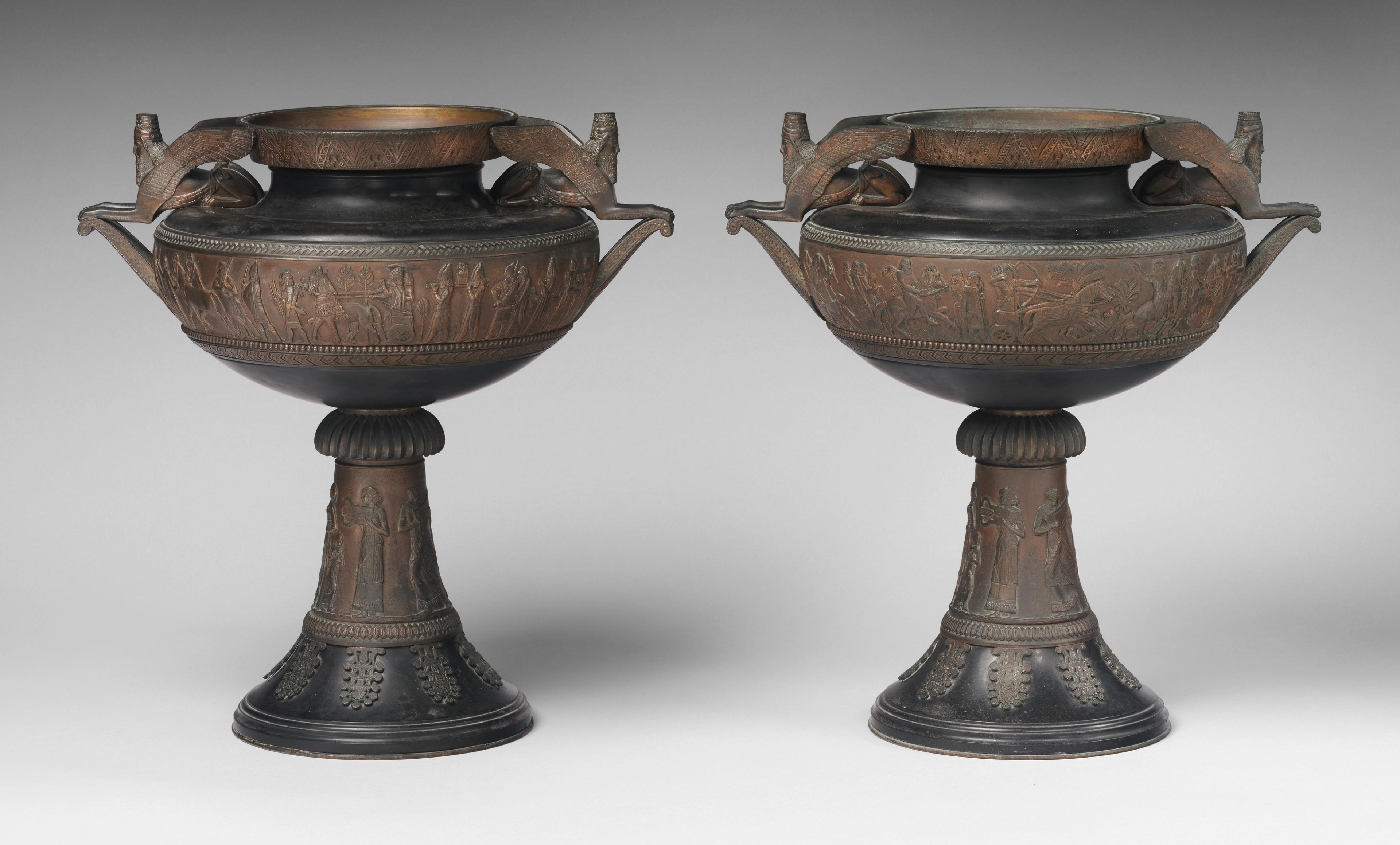 Photograph of two bronze urns with detailed Assyrian scenes on the bowl and stem