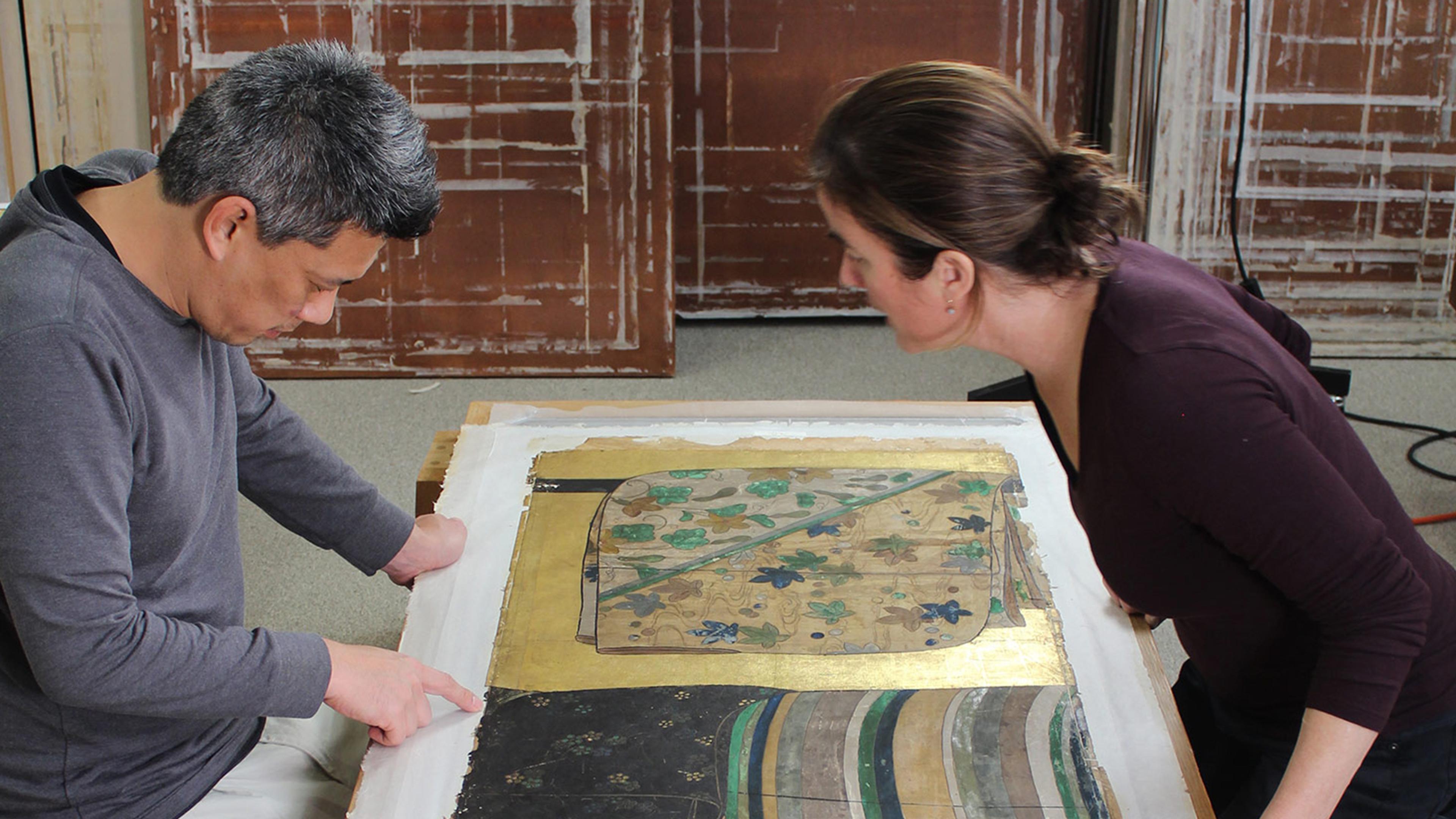 Two conservators kneeling over golden folding screens