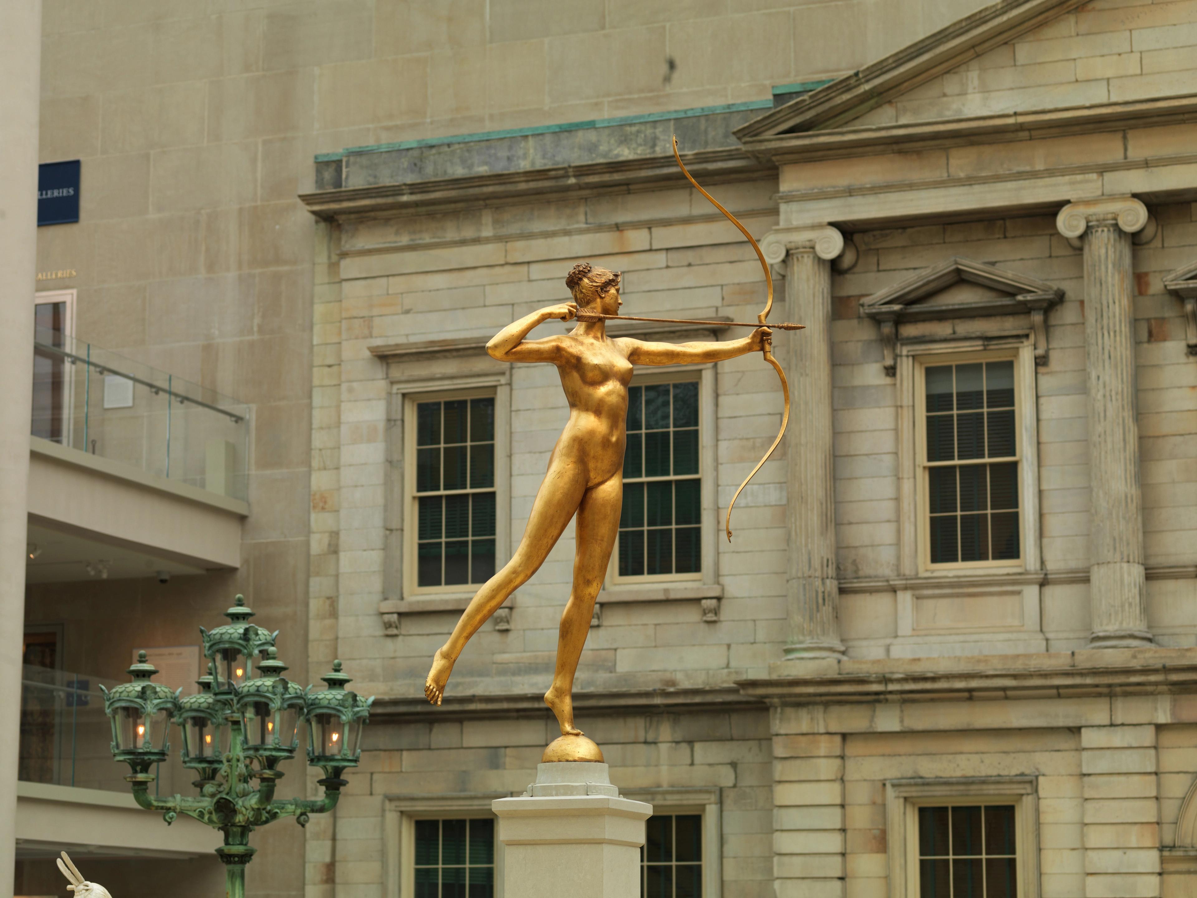 In gallery photo of a bronze sculpture of Diana, drawing a bow and arrow against a stoic building facade.