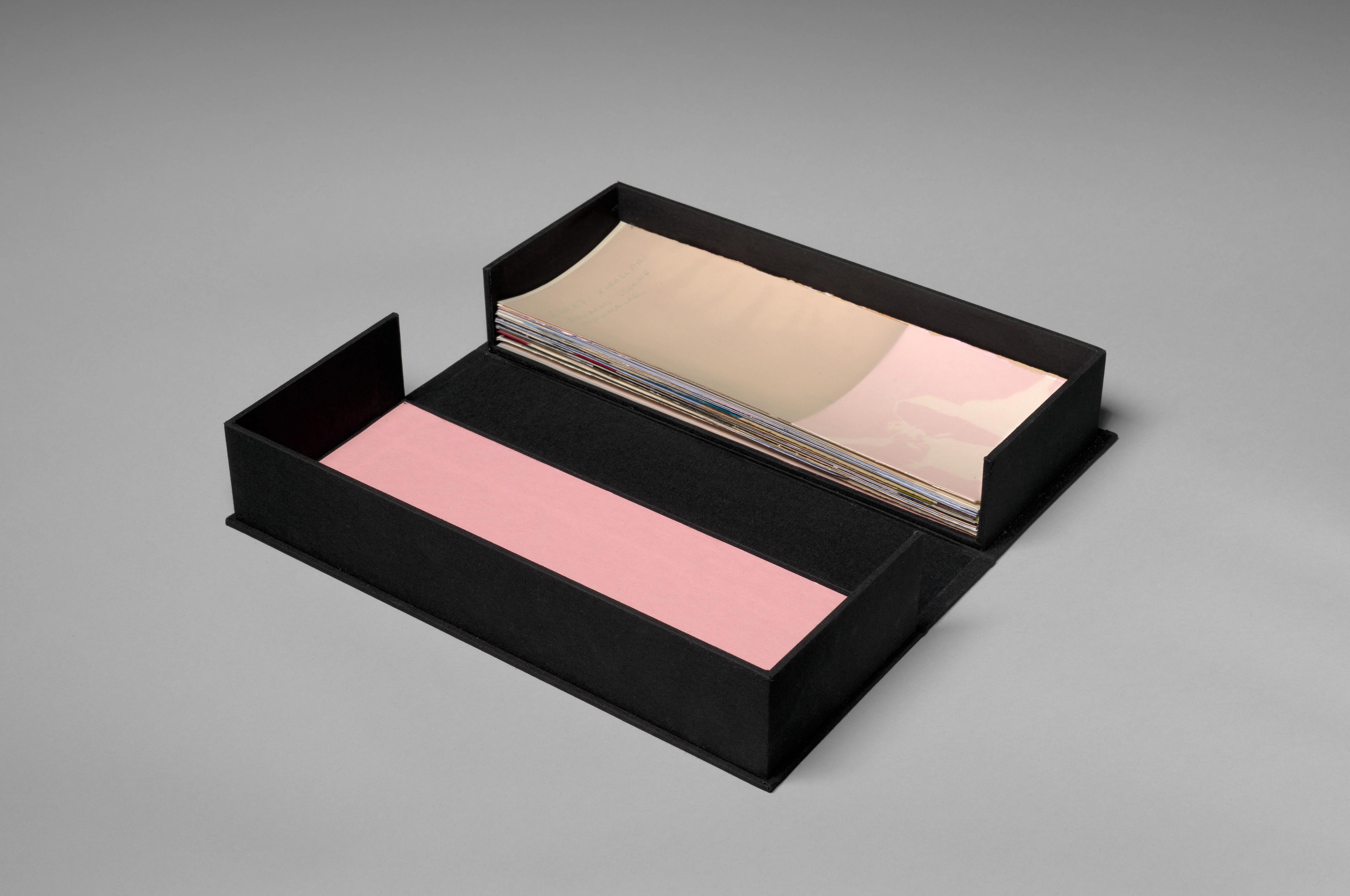 A small black box is opened and laid flat, holding small slips of paper in pinks and browns