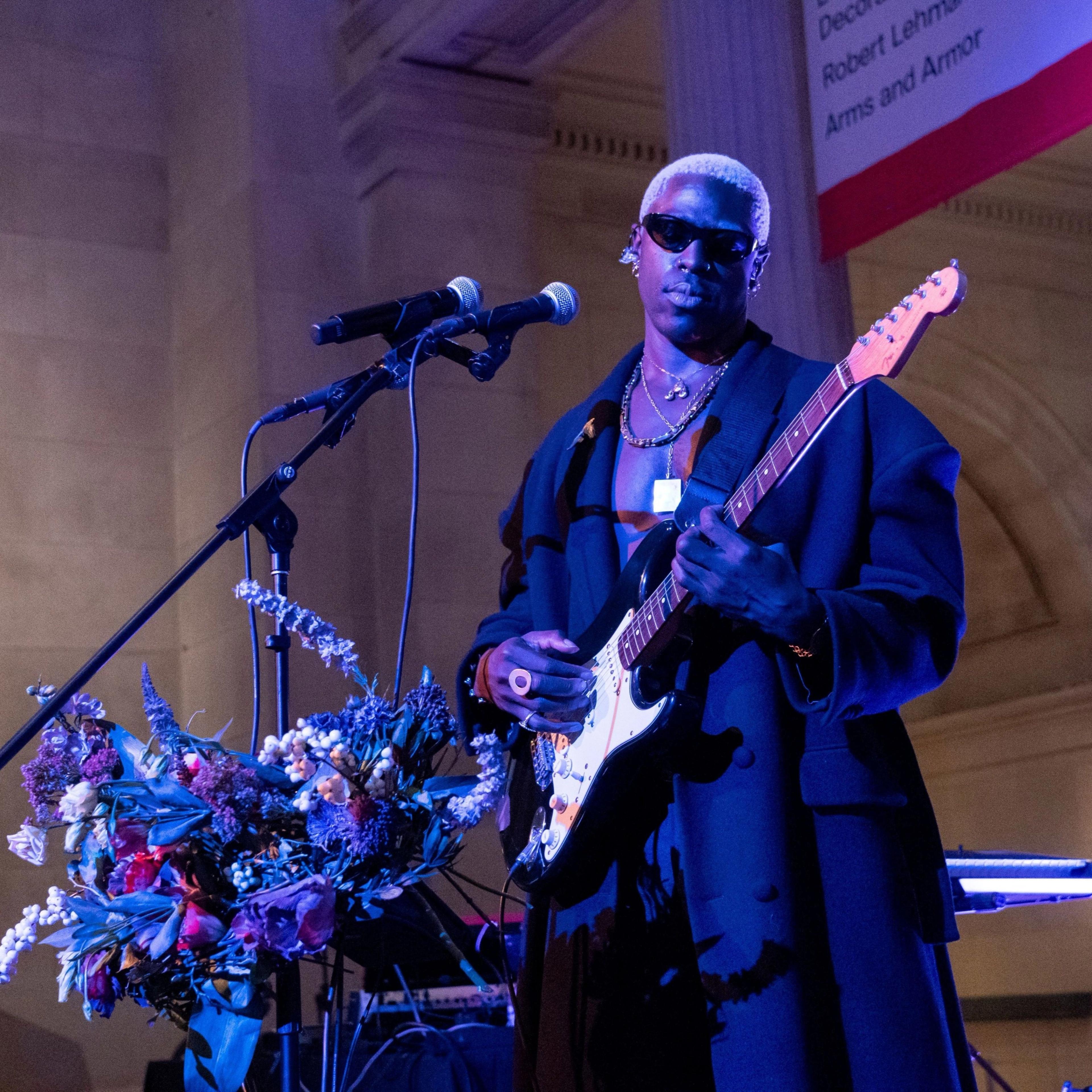 A singer wearing sunglasses, performing with a microphone and guitar in the Great Hall of The Met, with a column, light fixtures, and beige-colored stone walls in the background.