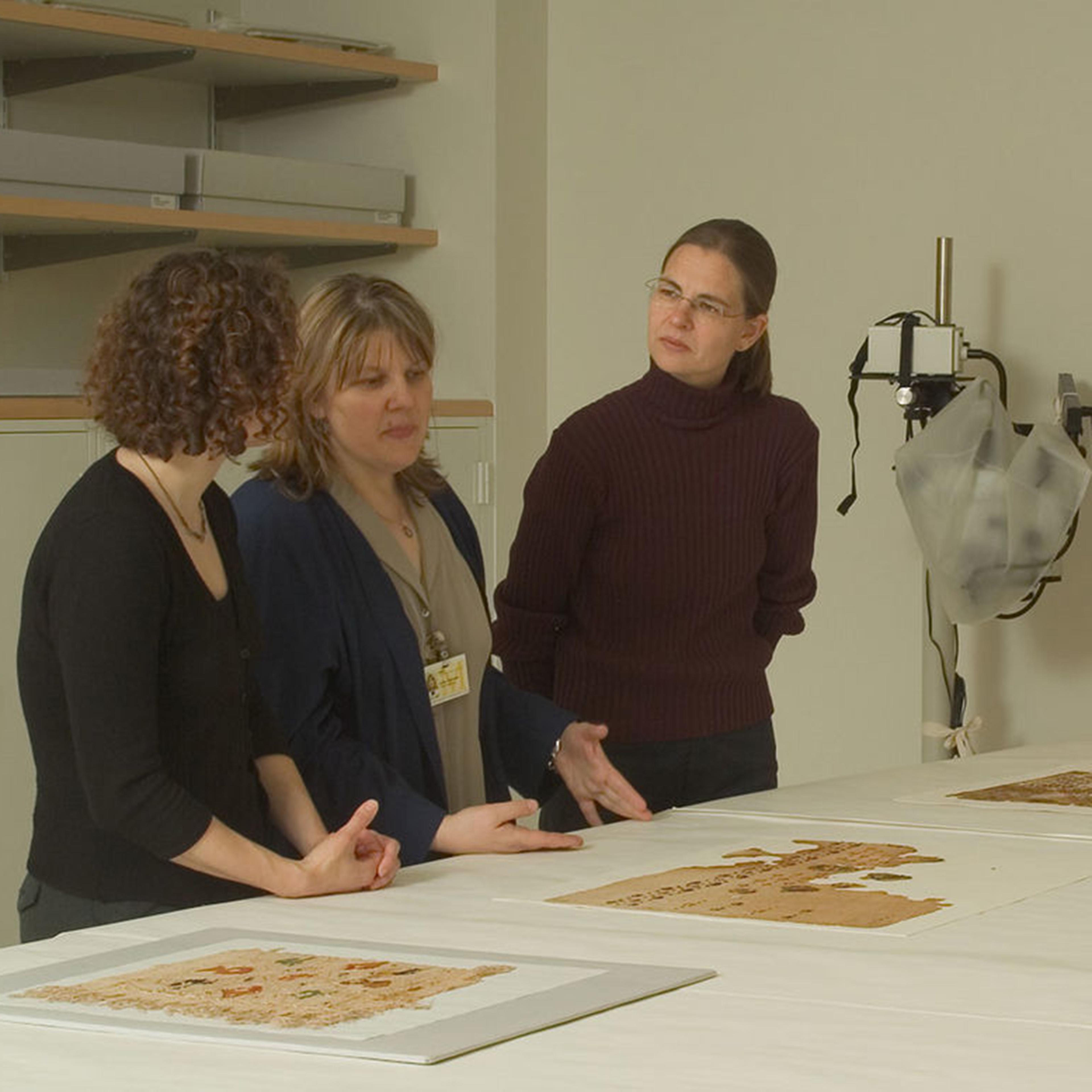 Three individuals discuss textile pieces displayed on a table in a conservation lab with a computer setup and other equipment in the background.