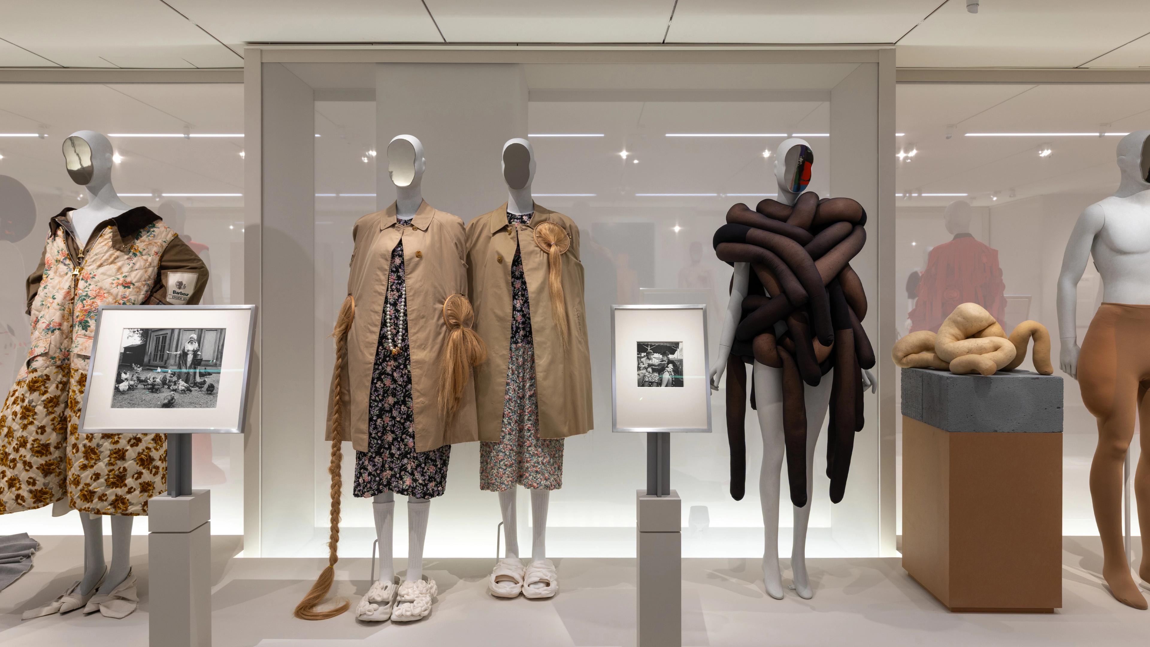 Mannequins in a modern gallery wear eclectic outfits including floral and abstract designs. Framed photos and artful fabric shapes 