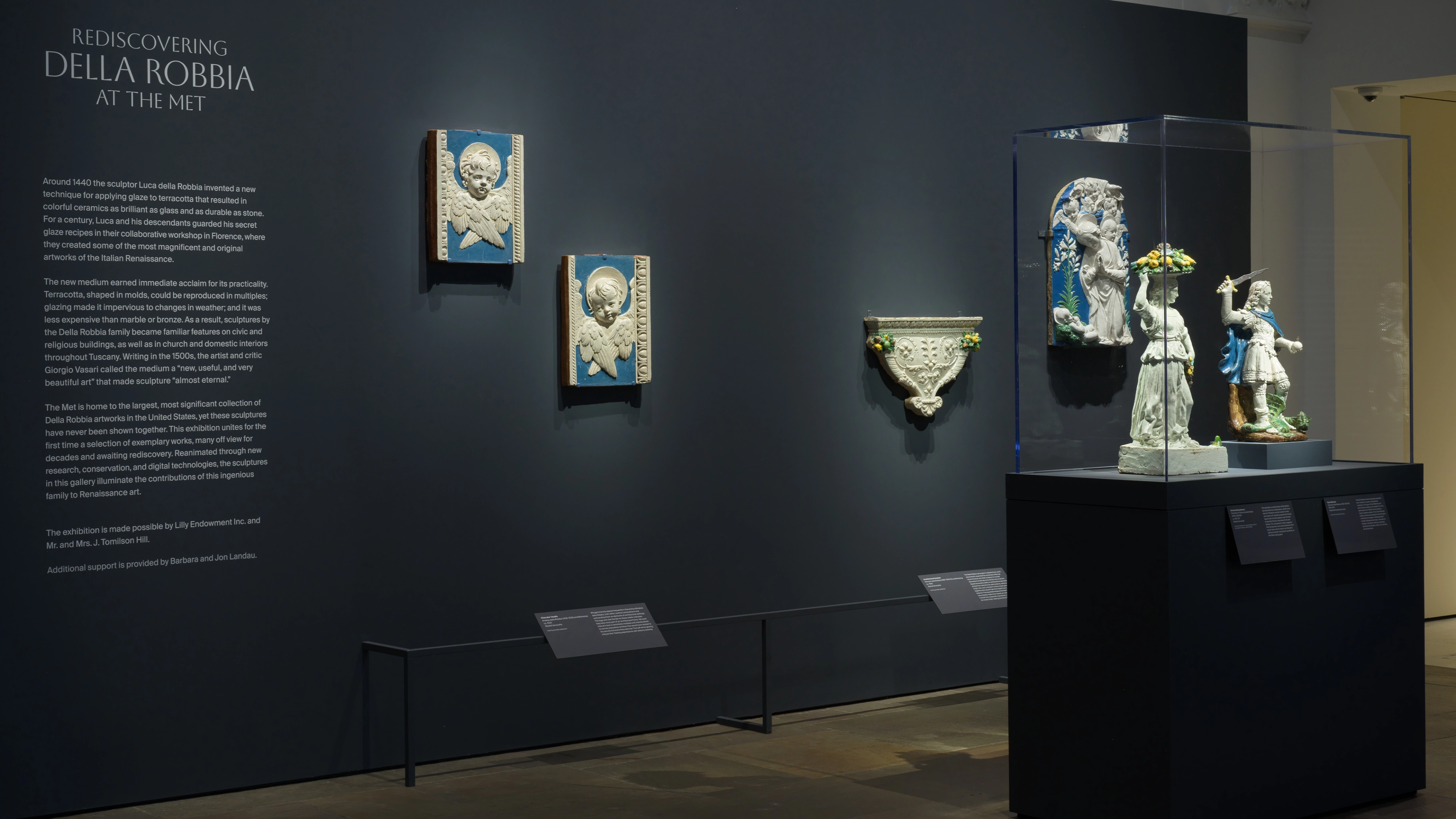 Dark museum gallery displaying blue-and-white Della Robbia ceramics: framed reliefs, a wall-mounted bowl, and figures in a glass case.