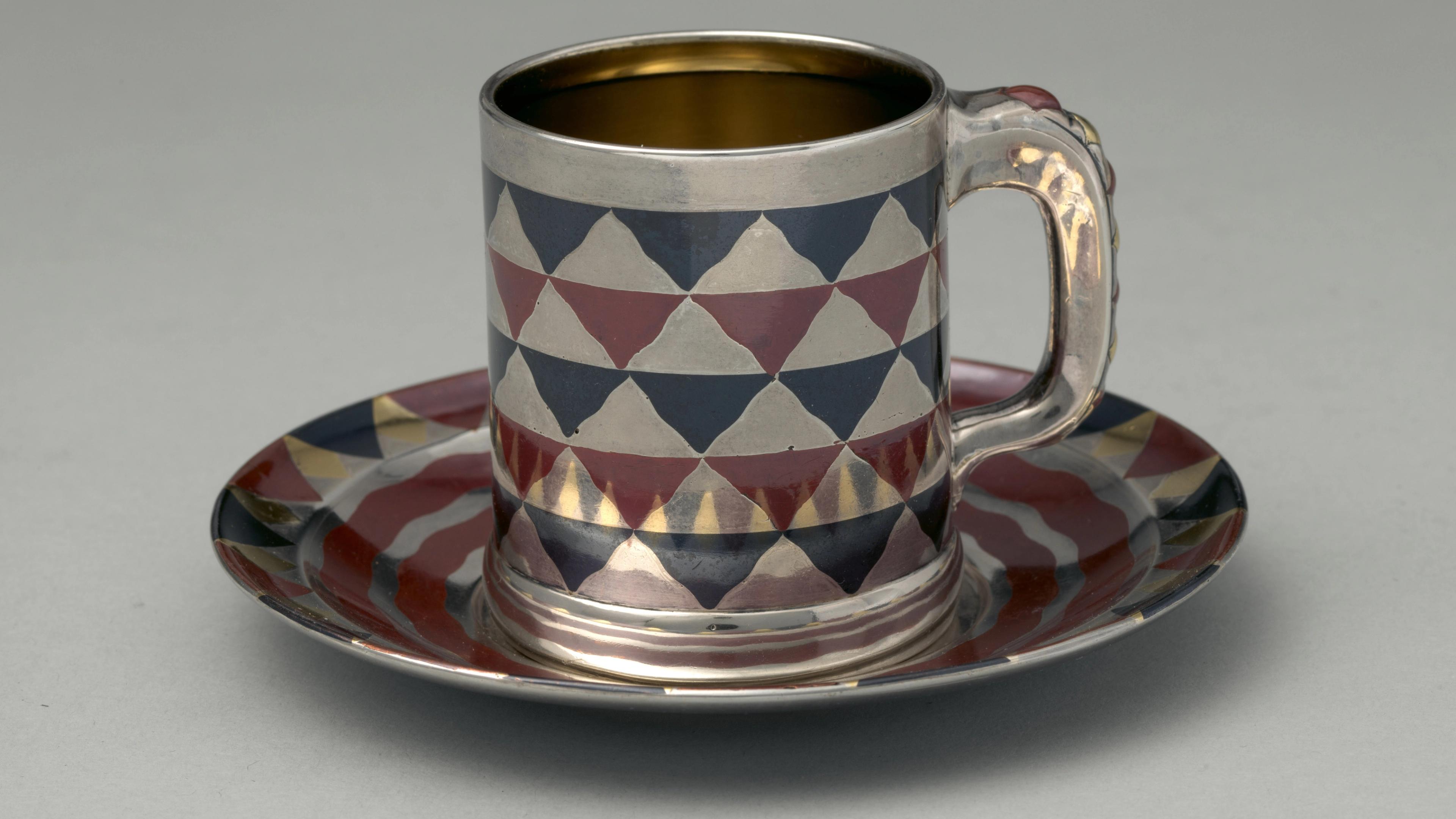 A silver, blue, and maroon cylindrical cup with a handle rests on a matching saucer. The cup and saucer are reflective and adorned with an inlay design of a checkerboard pattern of wavy triangle shapes.