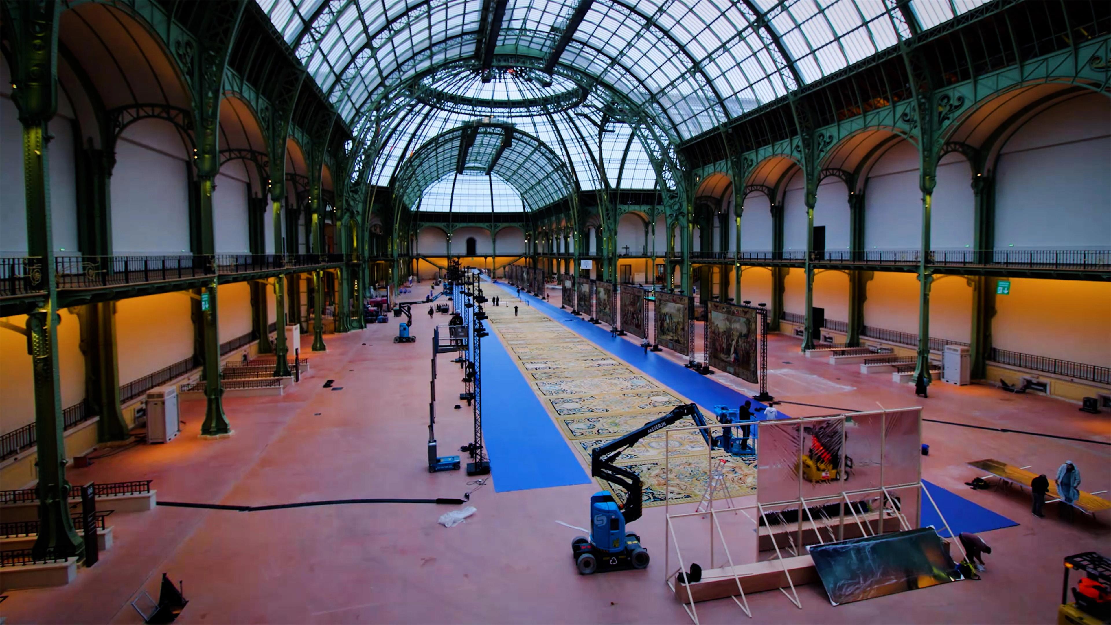 Grand glass-roofed historic hall with ornate iron arches, blue carpet, and exhibition displays; workers and lifts prepare the exhibits carpets for display.