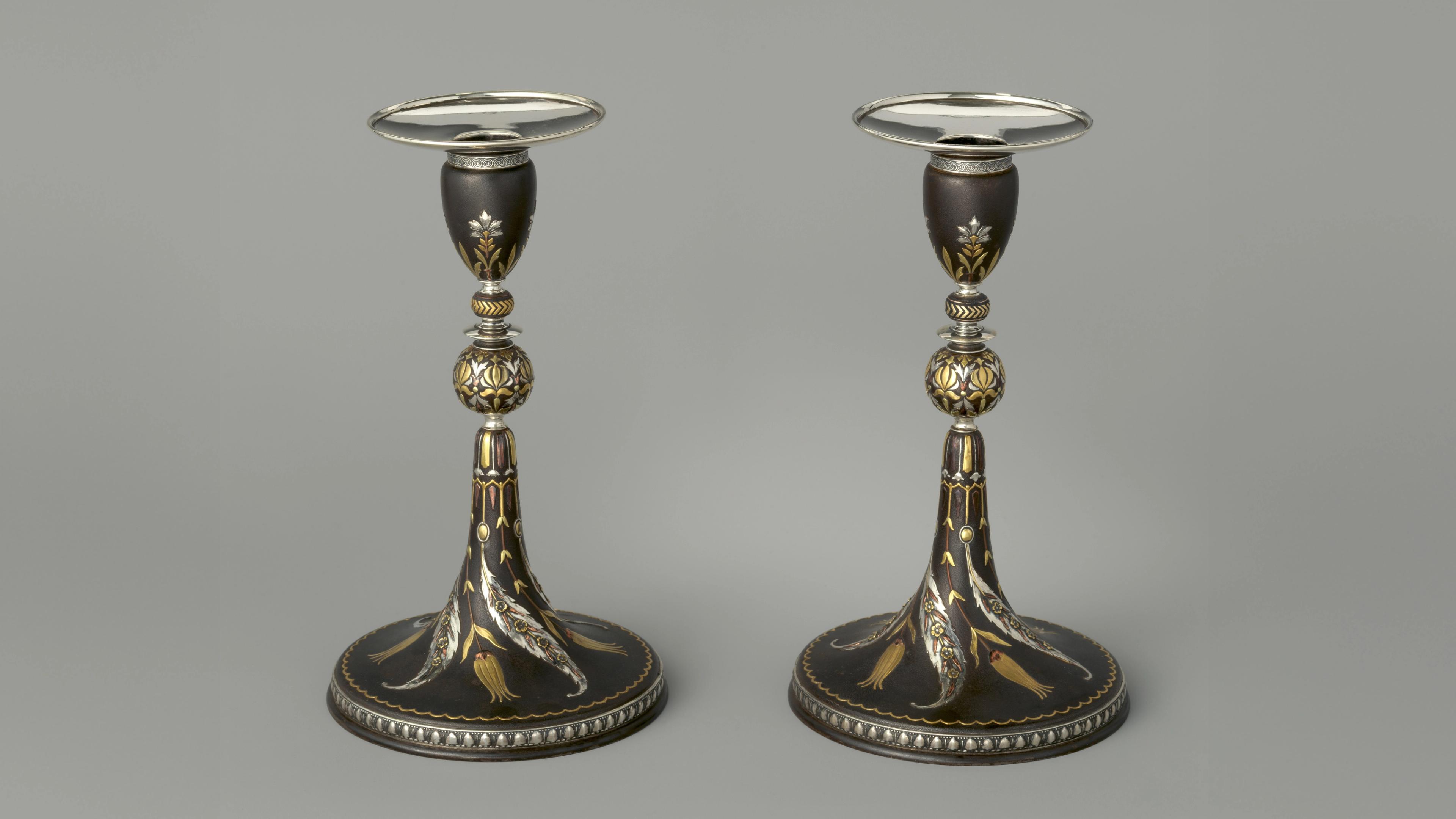 A pair of black iron candlesticks with a tapered conical base appear side-by-side. They are adorned with various sinuous floral motives that are modeled in low relief on the surface and surfaced in a variety of silver-, gold-, and copper-toned metals.