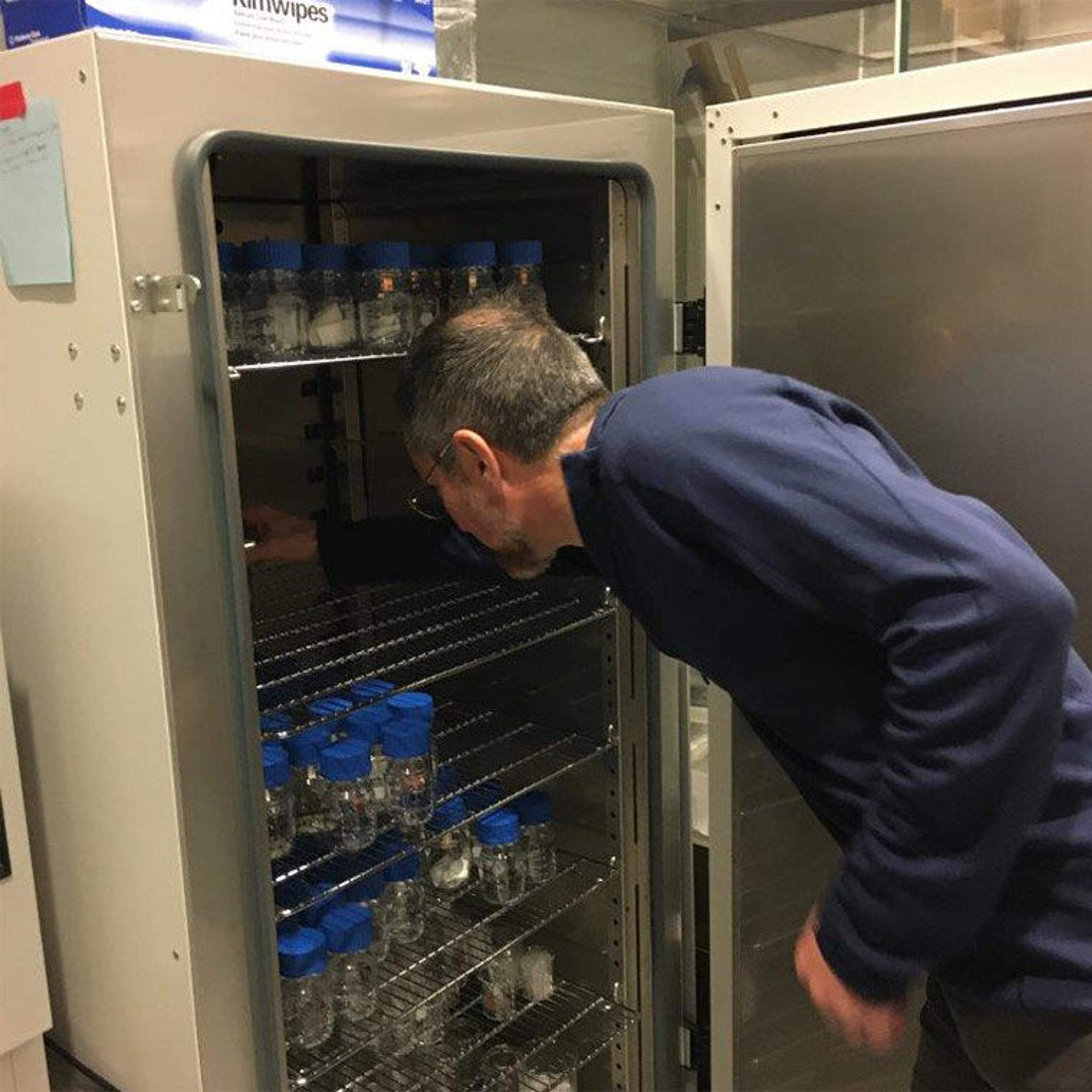 A man in blue coveralls reaches into a metal appliance full of test tubes in a laboratory.