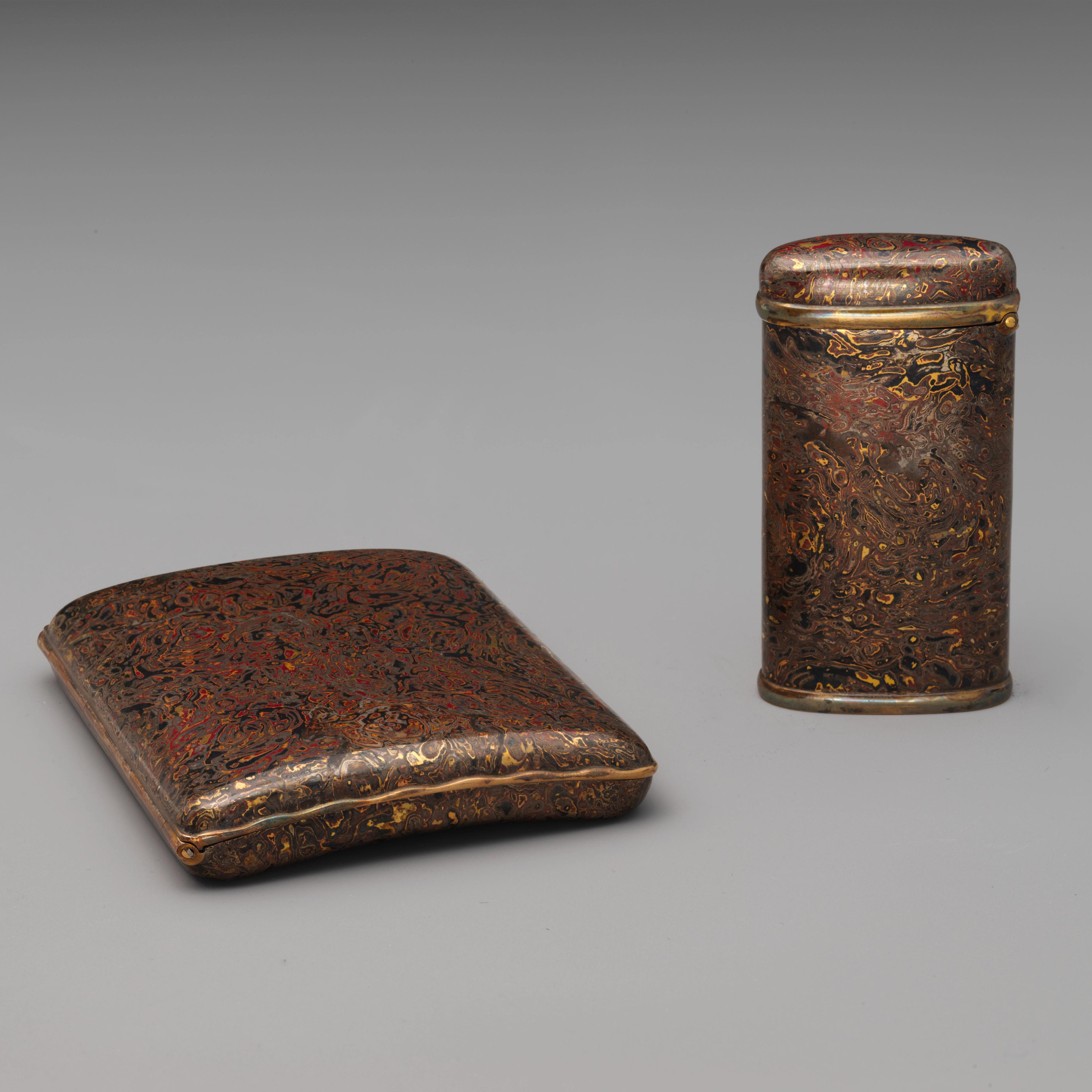 A flat cigarette case and a tall match case in a matching wood grain pattern rendered in deep red, black, brown, and gold metals. 