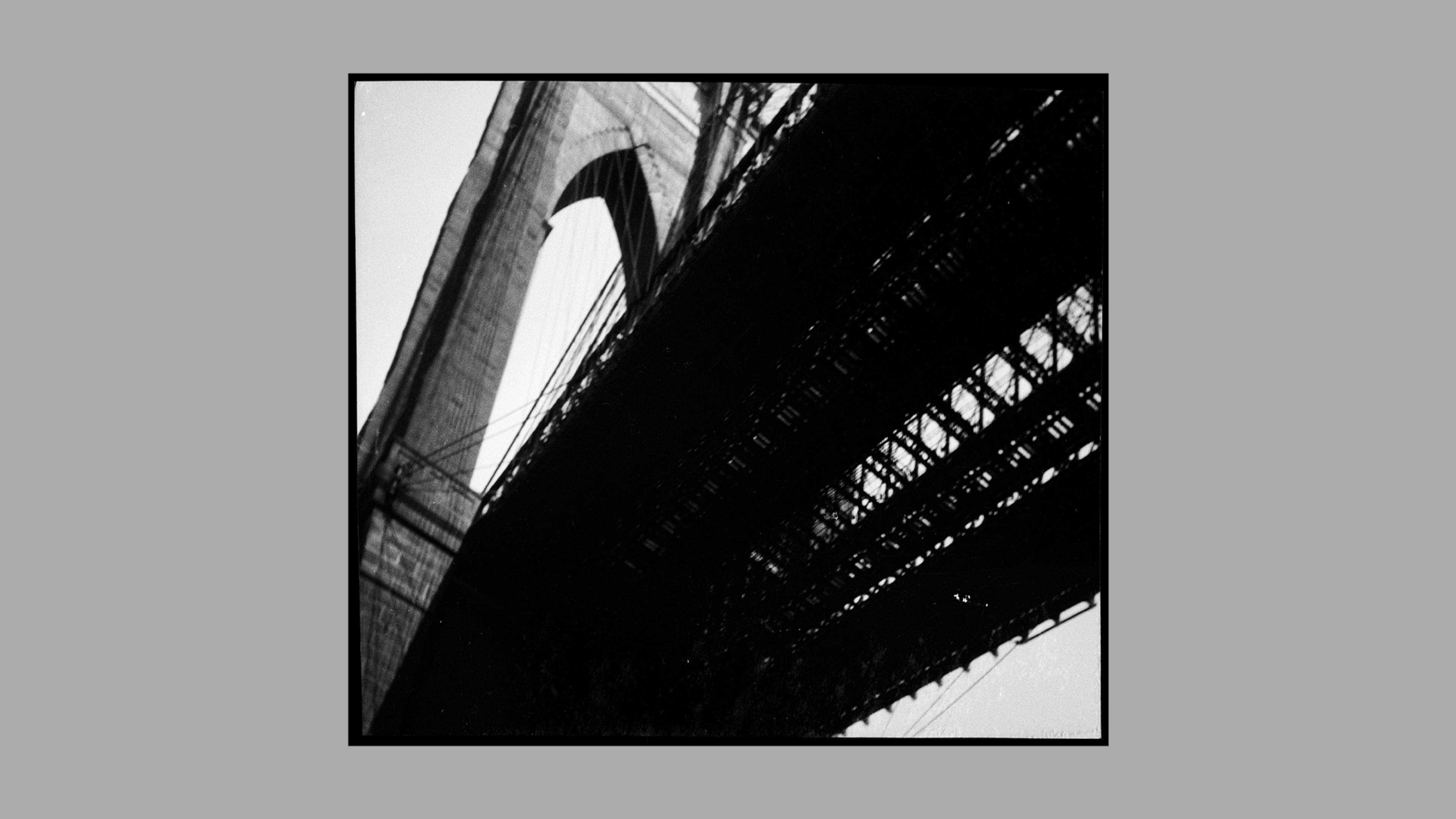 Square, black-and-white photograph of the underside of the Brooklyn Bridge, framed by rectangular gray matting.