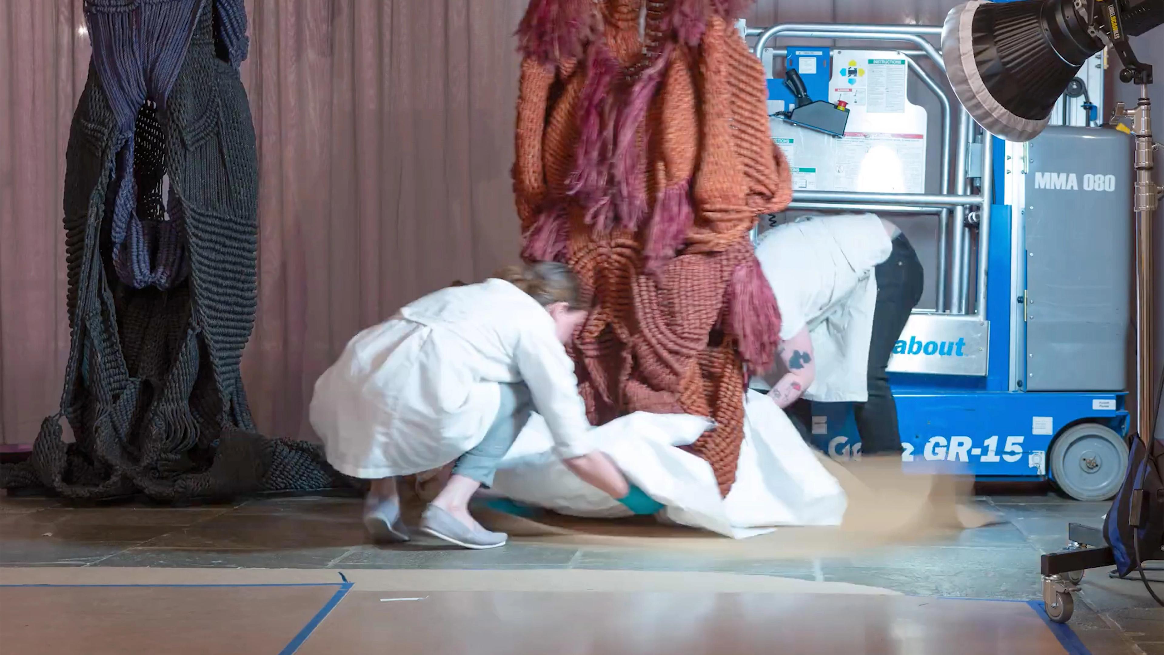 Two kneeling conservators adjust a shite sheet at the base of an orange and maroon ensemble.