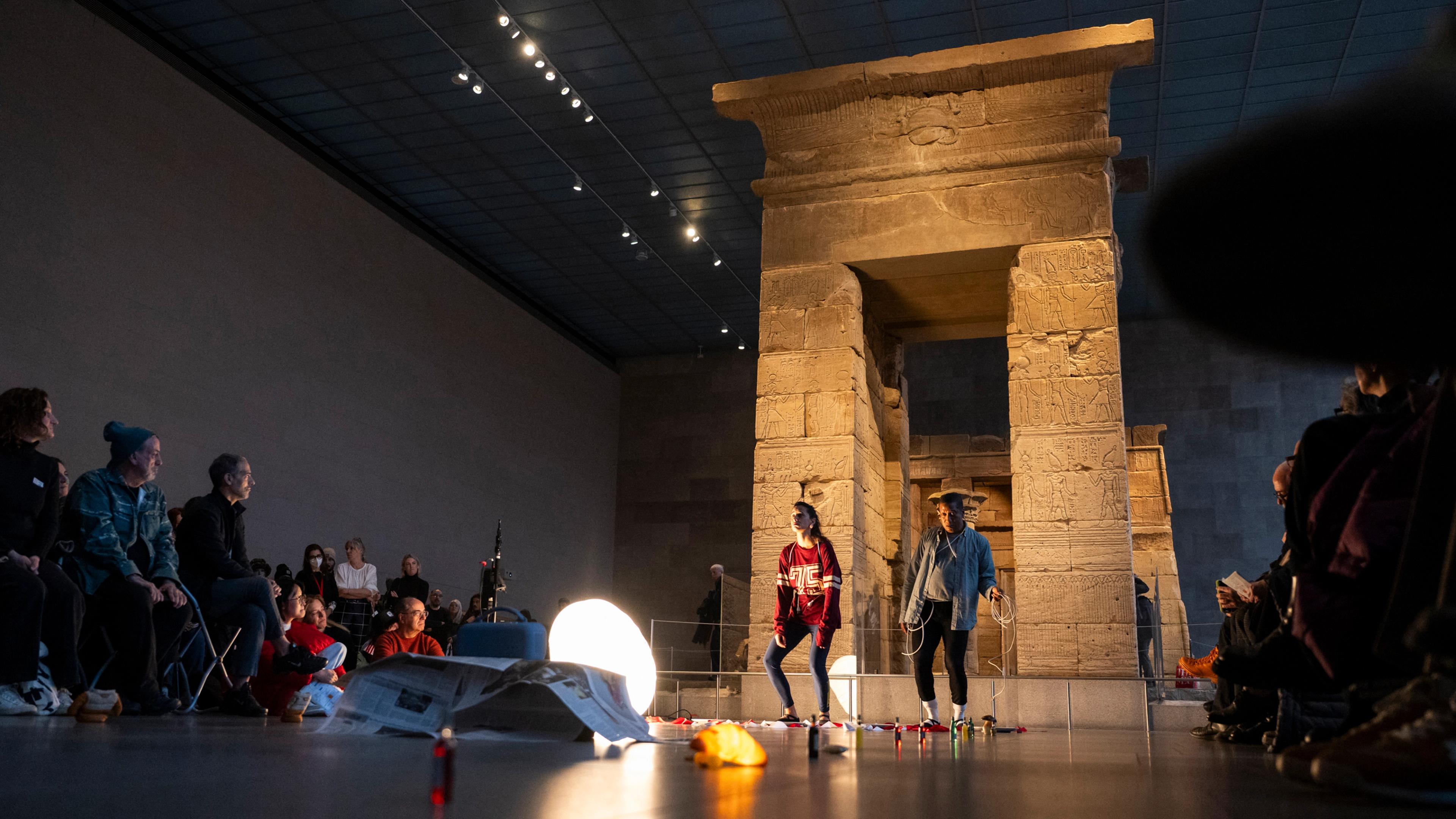 Visitors watch two performers in front of an ancient Egyptian temple installed in a gallery, with a glowing light and scattered objects on the floor.