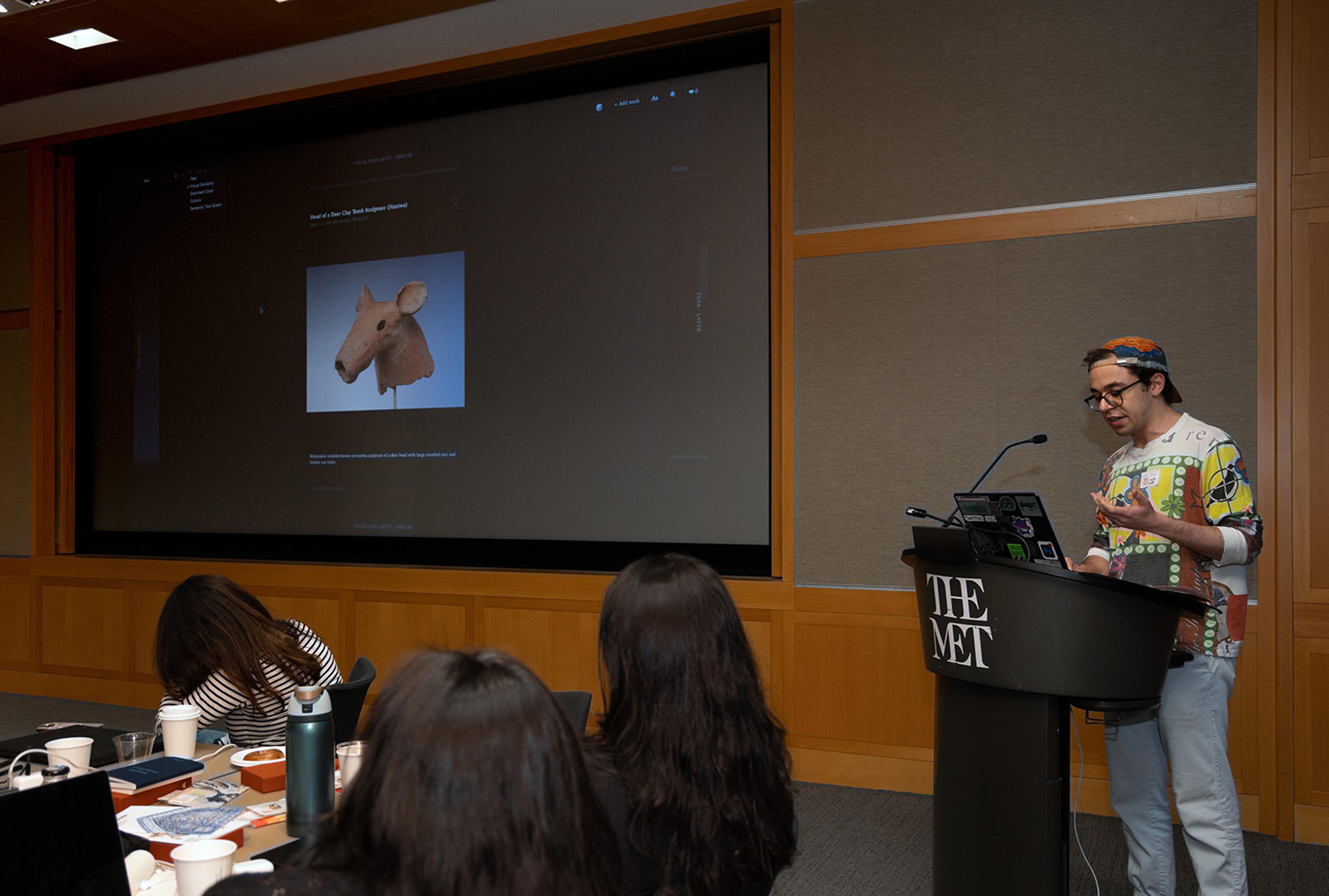 A presenter in a colorful sweater speaks at a podium marked "The Met," with a large screen displaying an animal head sculpture.