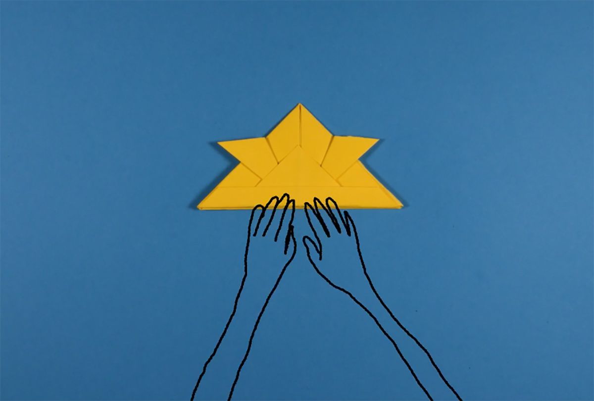 Animated hands work to fold a yellow oragami helmet placed on a blue background.