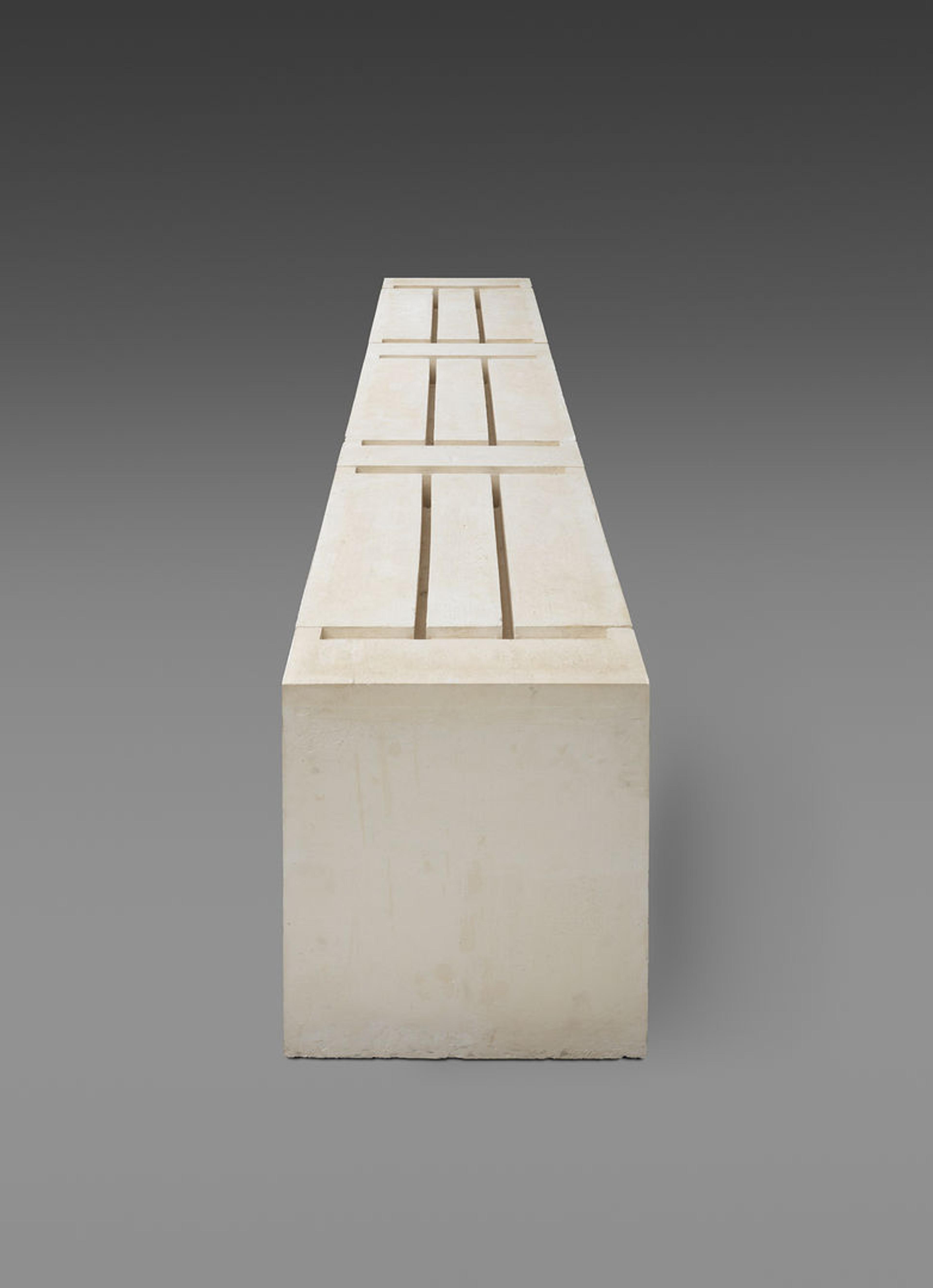 Bulky, rectangular plaster sculpture against a gray background. Grooves on the upper side of the sculpture outline the underside of three tables.