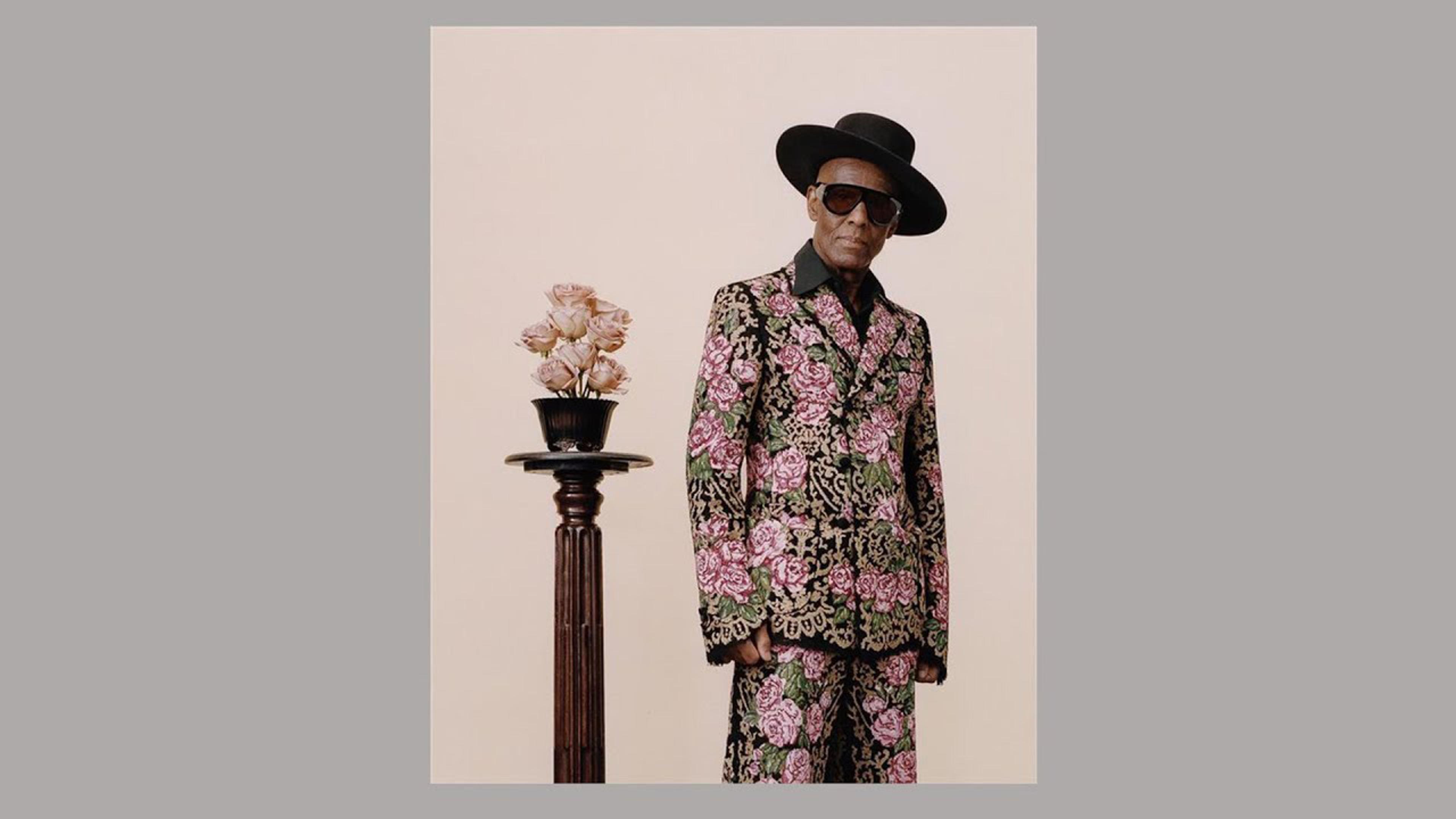 Older man dressed in a floral-printed suit, wide-brimmed black hat, and sunglasses poses next to a pedestal topped with flowers.