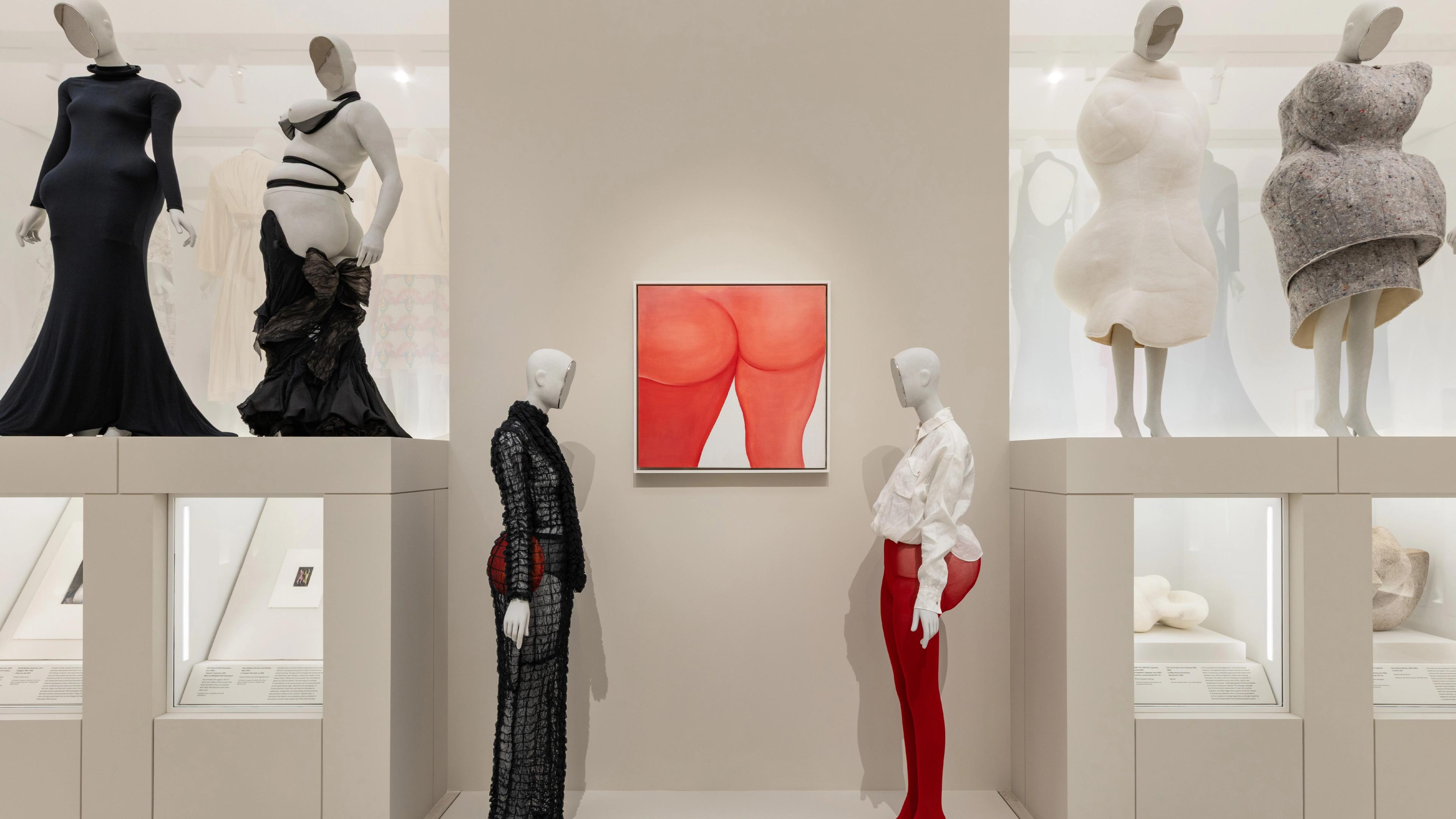 Exhibit of avant-garde fashion mannequins in creative, textured outfits on pedestals, with a central artwork depicting orange-red abstract shapes.