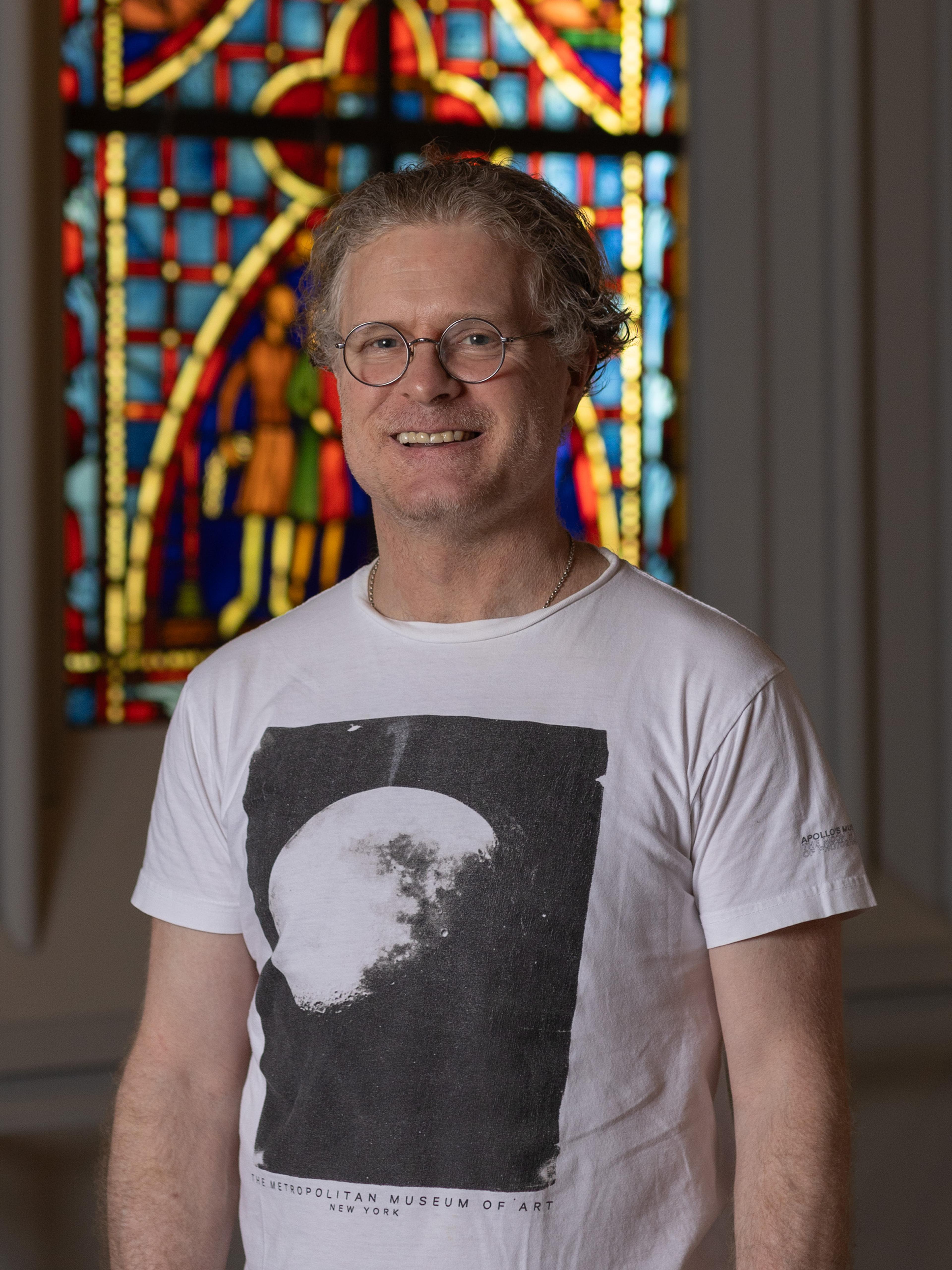 A man wearing a white t-shirt and wire glasses smiles in front of a stained glass window