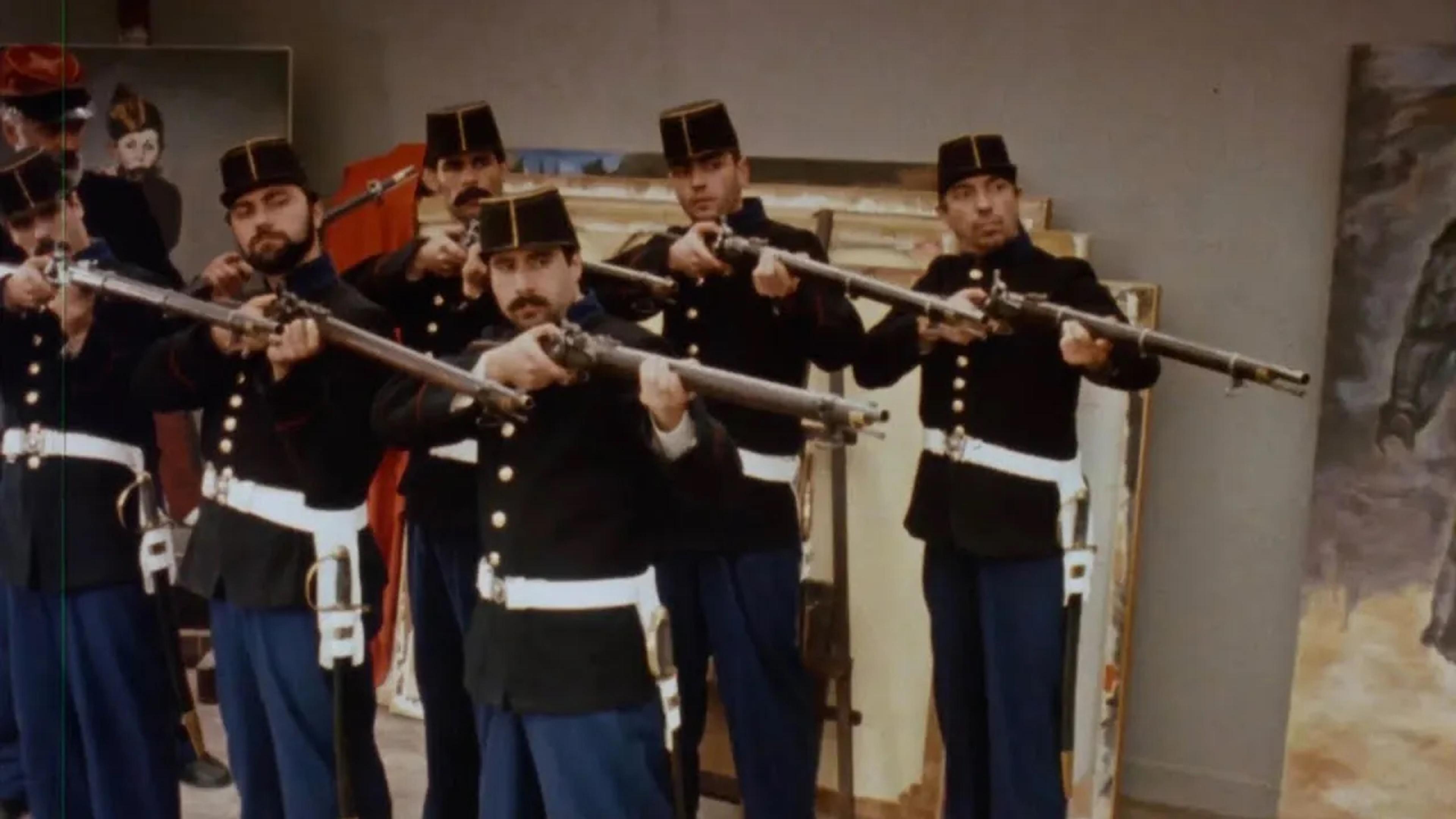 A group of men in historical military uniforms aim rifles in an art studio with paintings in the background.