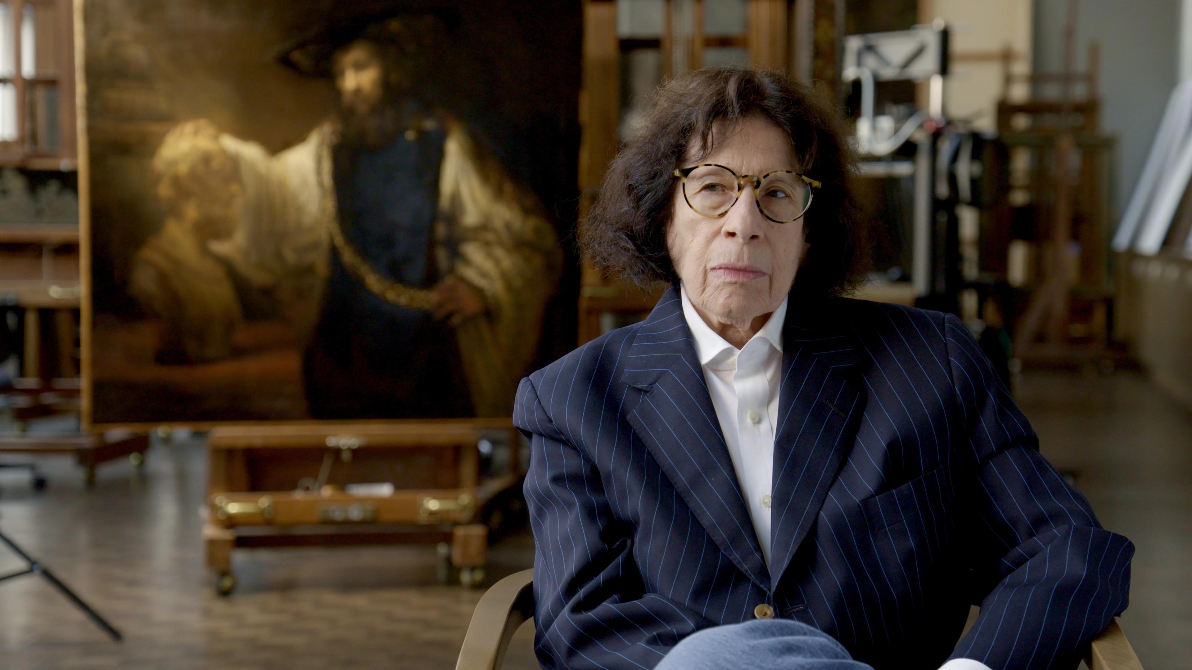 Writer Fran Lebowitz sits in The Met’s Paintings Conservation Lab in front of Rembrandt’s Aristotle with a Bust of Homer painting.