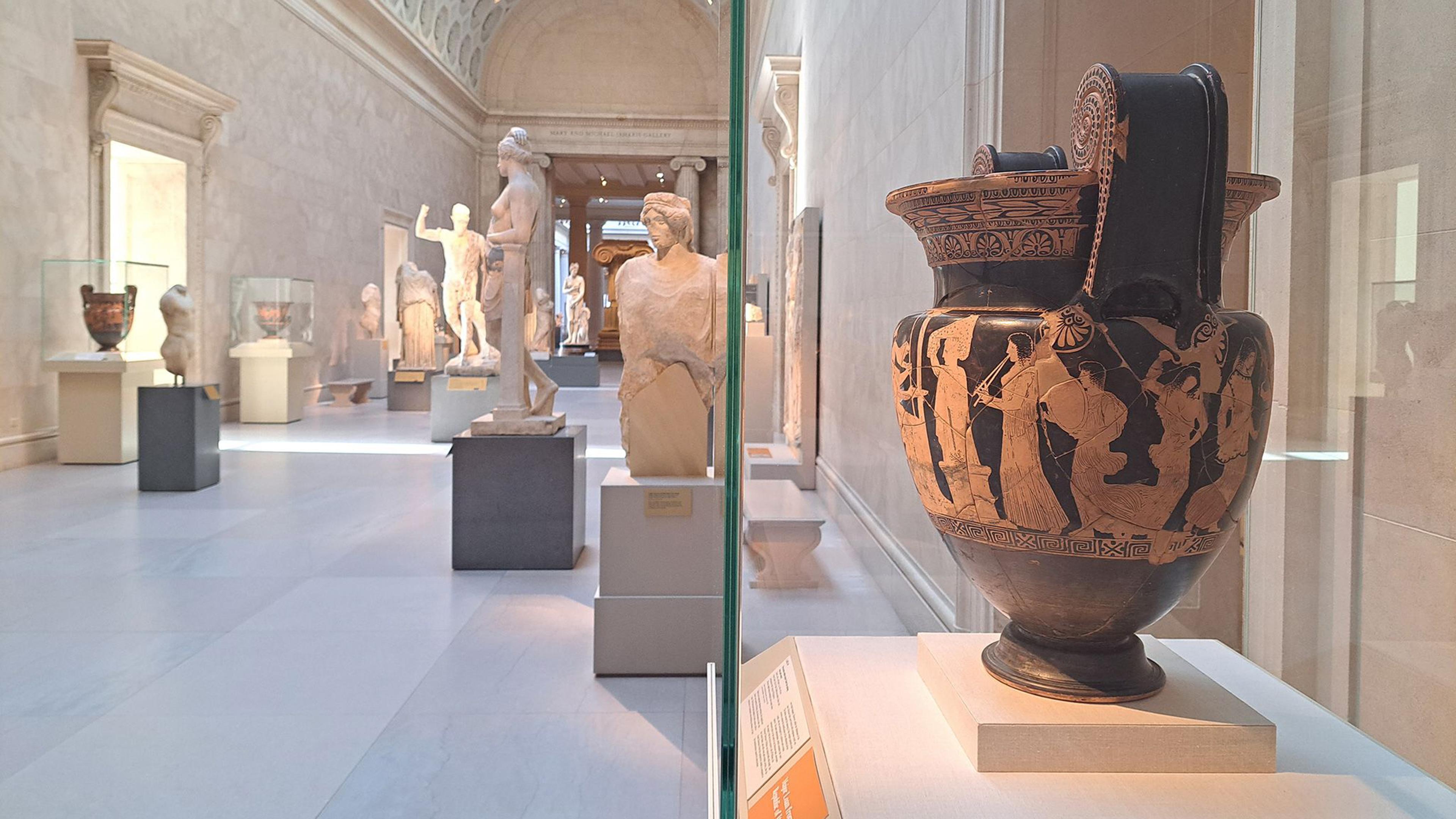 In the foreground at right, side view of a red and black krater in a glass case. In the background, the sunlit hall of the Greek and Roman galleries, with numerous statues and other glass cases in the distance