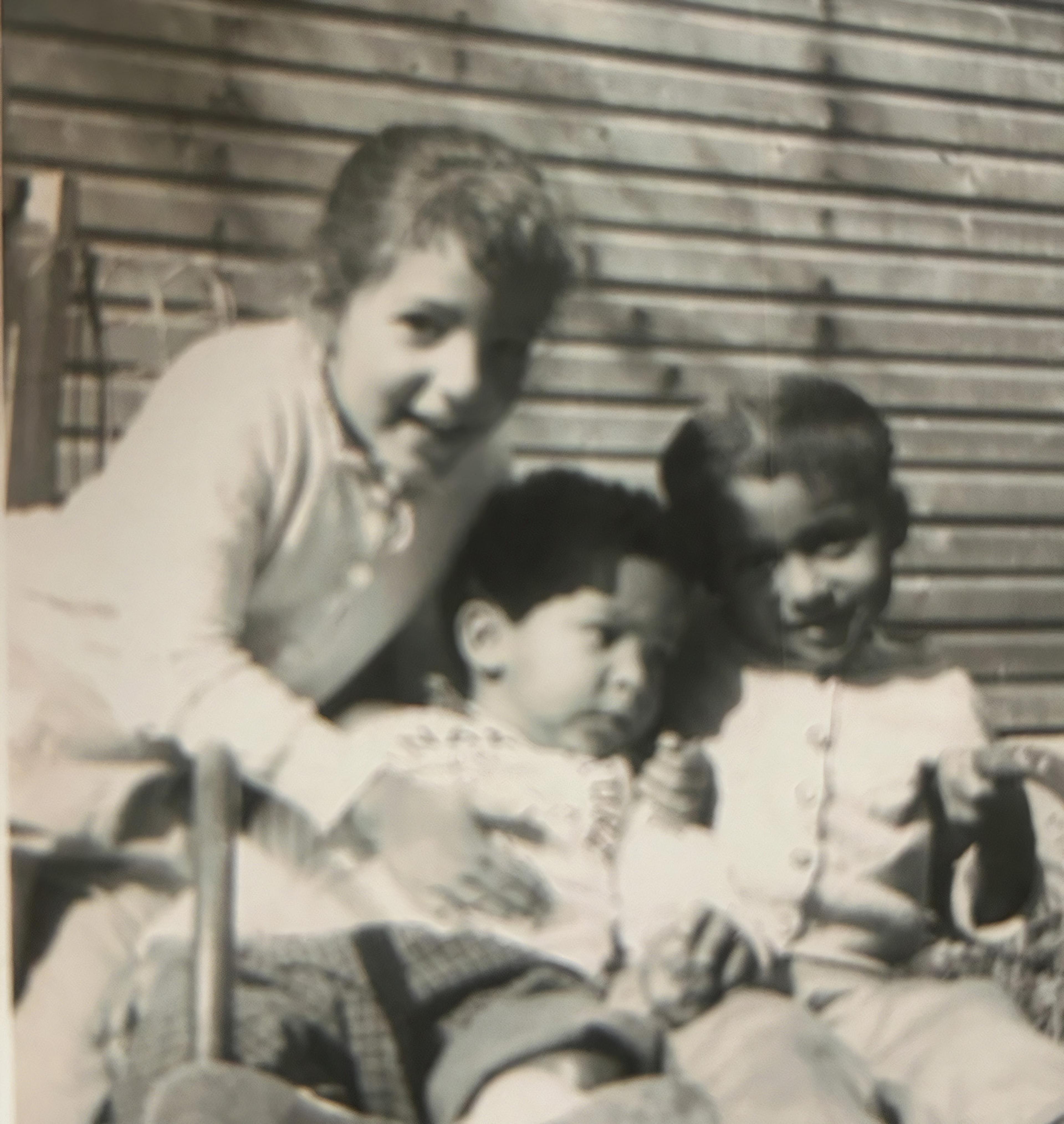 A vintage black-and-white photo of three smiling children, two girls and one boy, sitting closely together on a bench against a wooden wall.