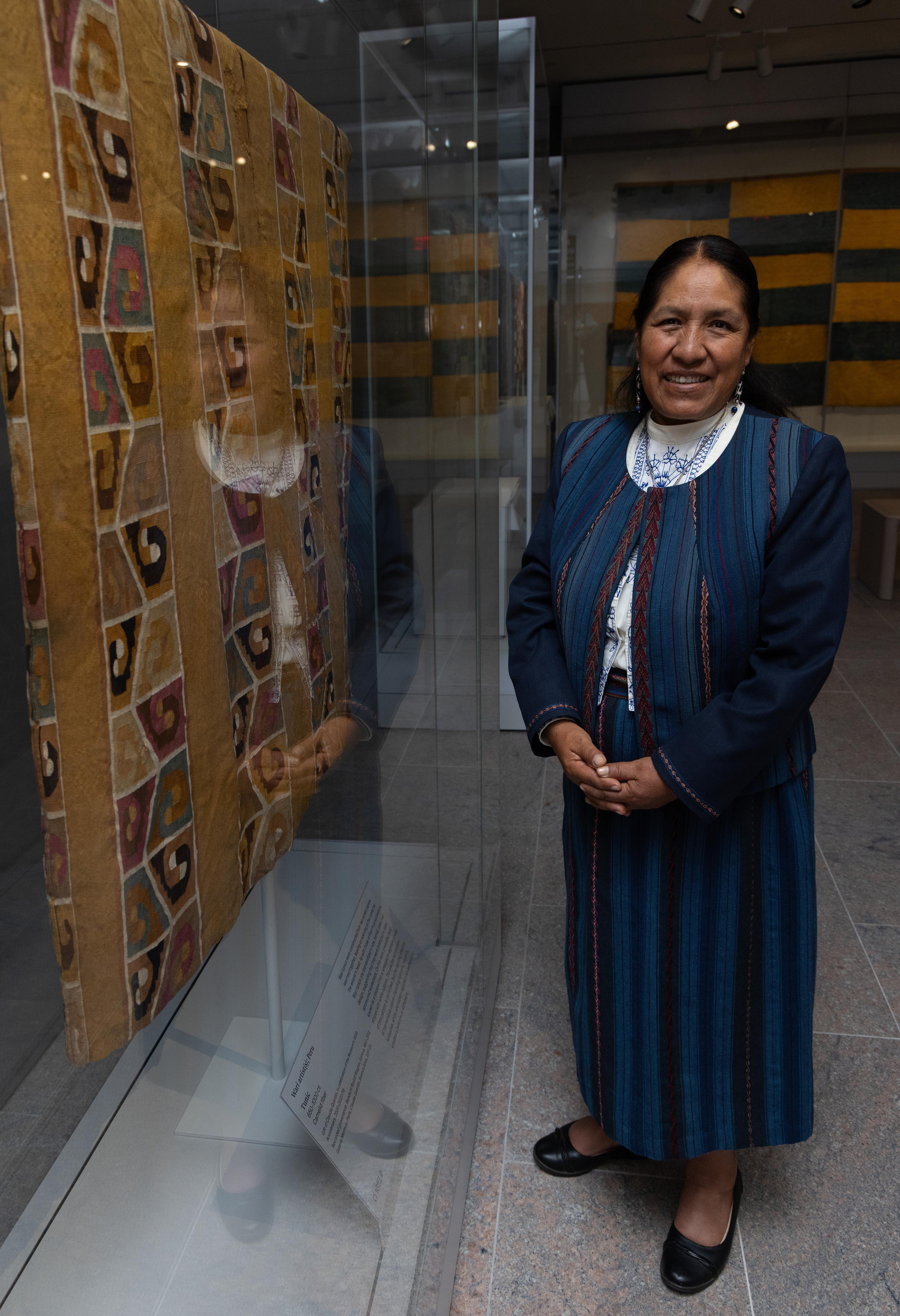 Nilda Callañaupa Alvarez smiling and standing in front of a glass case with a tapestry tunic inside