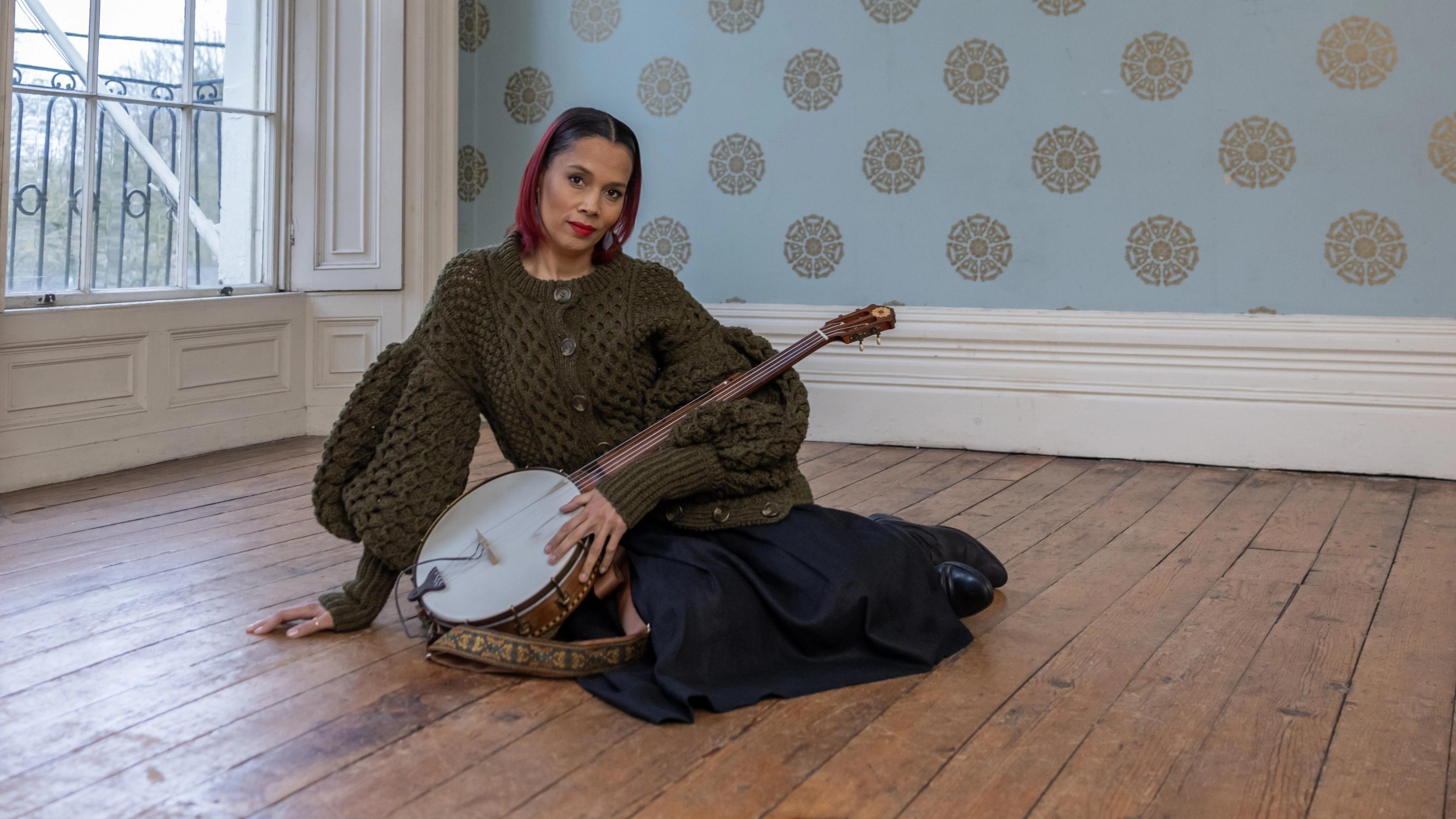 A woman in a dark green sweater and skirt poses sitting on a hardwood floor with a banjo.